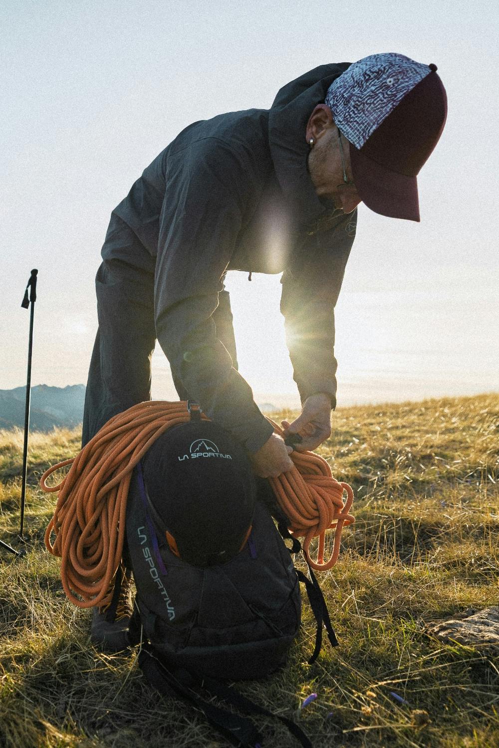 A mountain athlete adjusting his gear and climbing rope on a grassy ridge, featuring La Sportiva trail running and mountain equipment