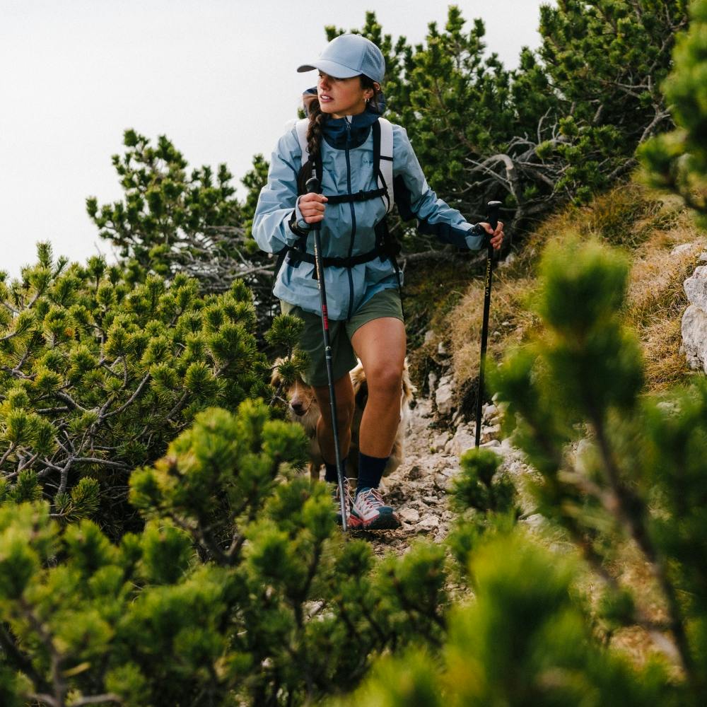 A woman hiker with trekking poles navigating a lush green mountain trail in La Sportiva women's hiking apparel and footwear.