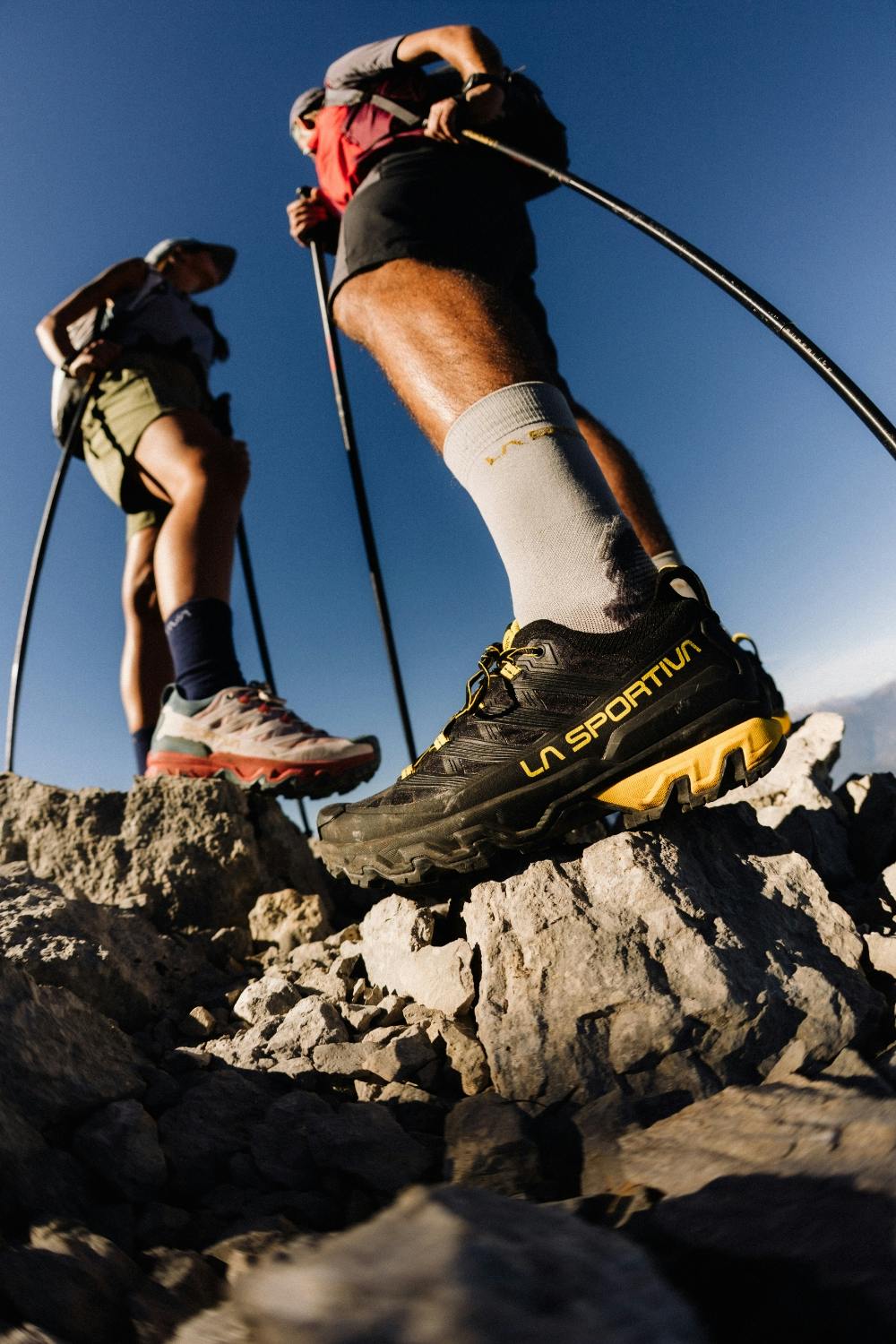 Close-up of La Sportiva hiking boots on a rocky mountain summit with hikers trekking in the background under a clear blue sky.