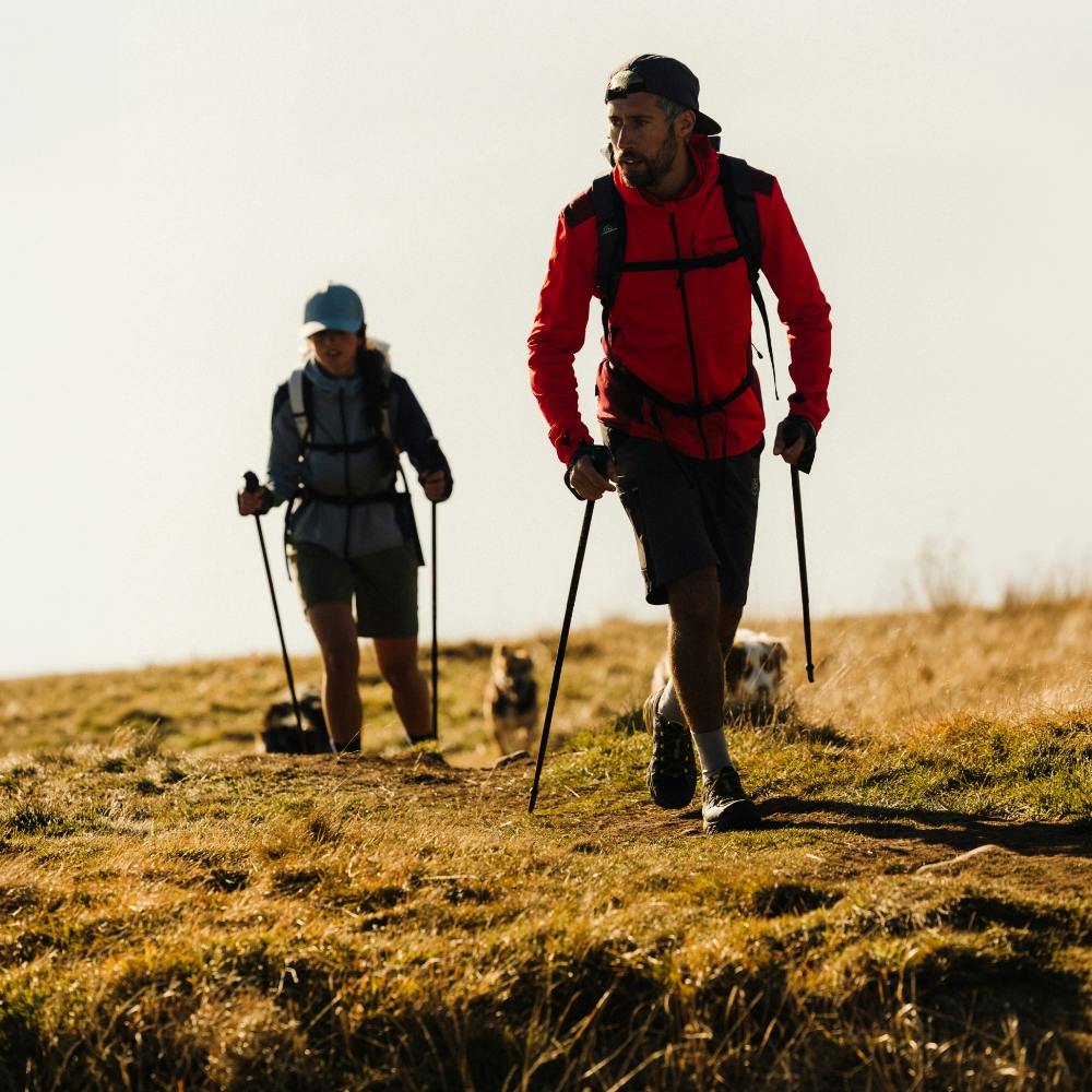 Two hikers in technical gear, including a man in a red jacket, trekking across an alpine meadow in La Sportiva men's outdoor clothing.