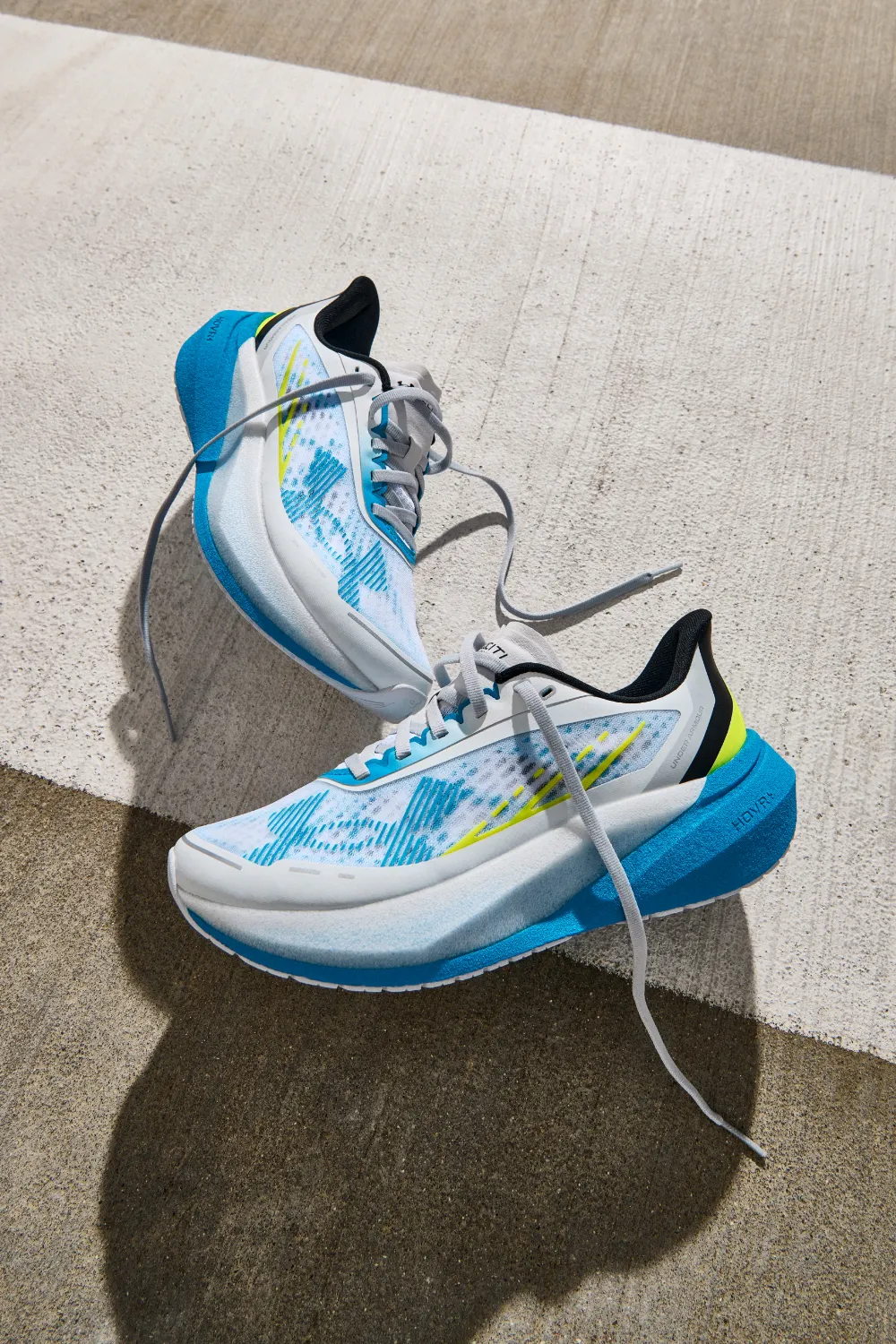 Pair of Under Armour blue and white marbled Velociti Elite 3 racing shoes sitting on a sunlit concrete surface.