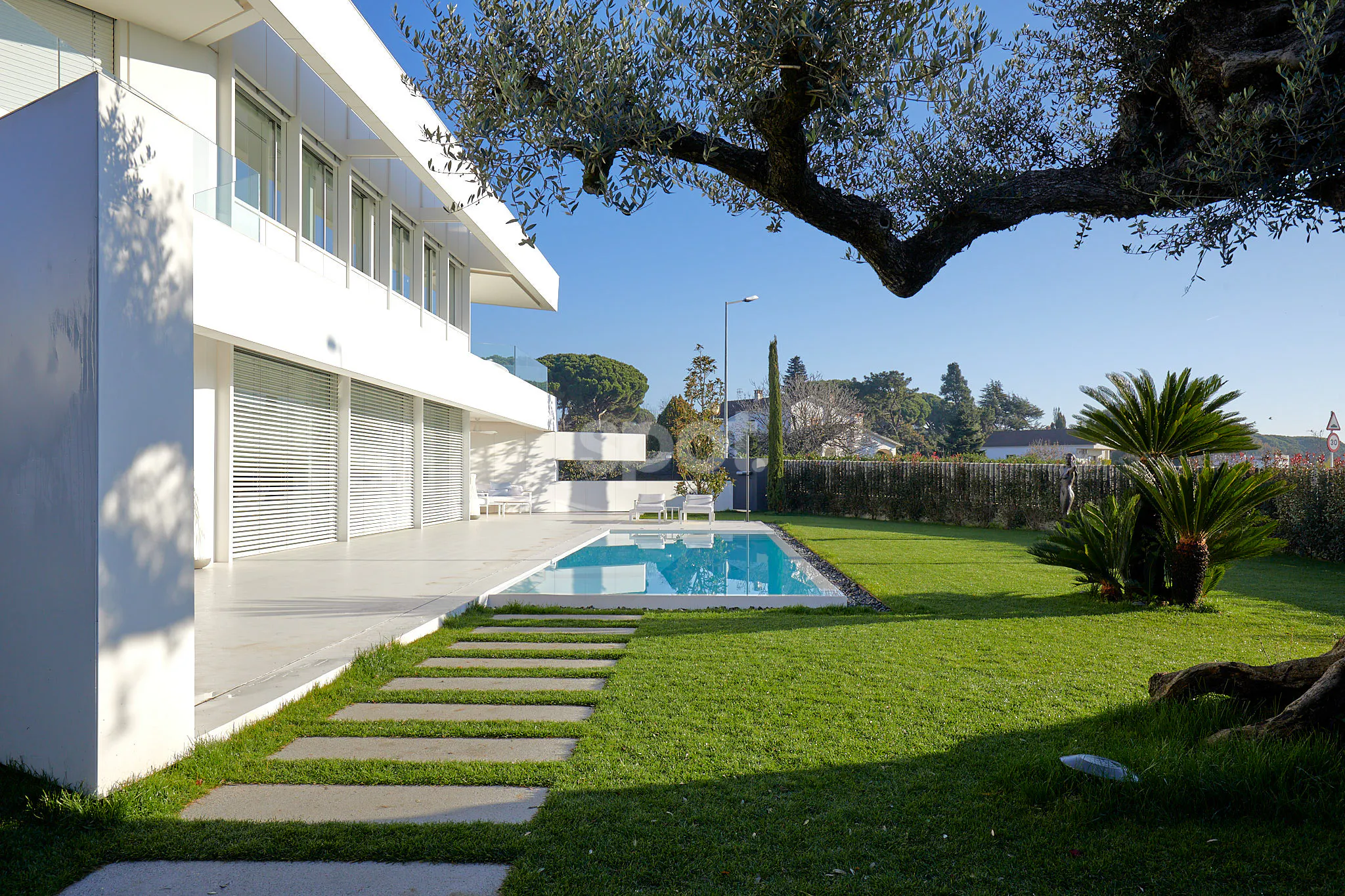 Localización de rodaje en una villa blanca moderna de España con piscina, jardín paisajístico, olivo, balcones de cristal y luz mediterránea.