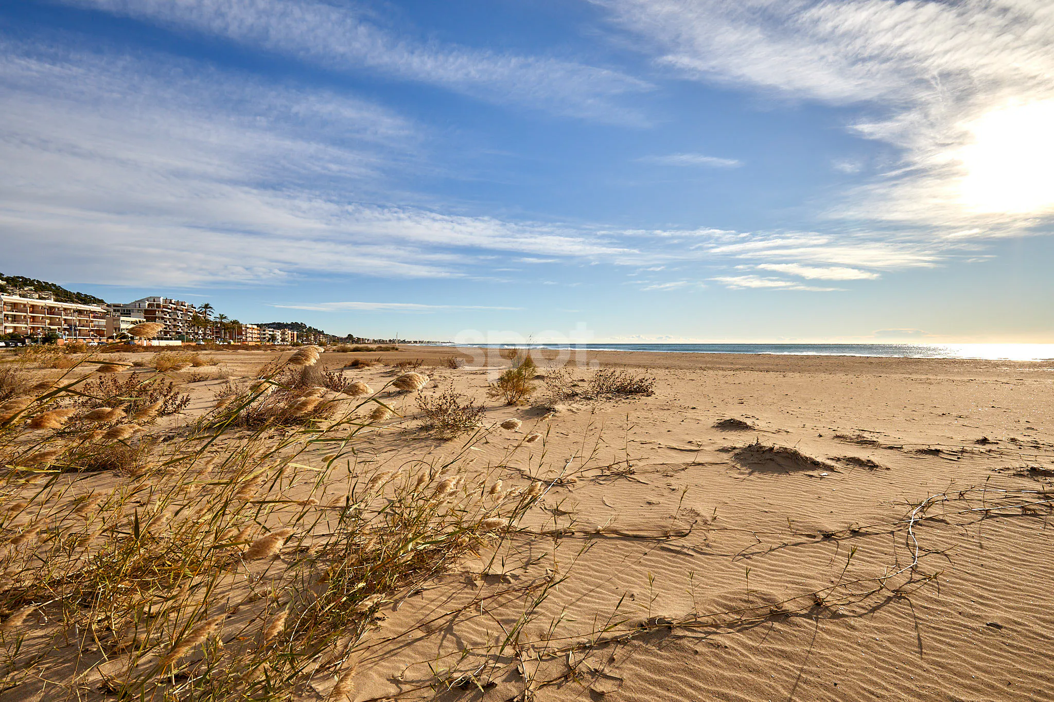 Mediterranean beach filming location in Mallorca with sand dunes, sea views, coastal buildings, blue sky and natural sunlight.