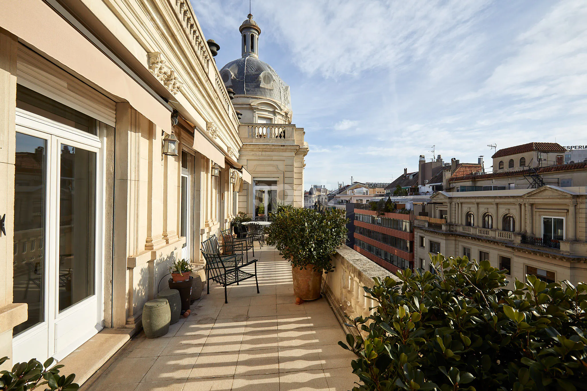 Elegant rooftop terrace filming location in Barcelona with classical architecture, a domed roof, outdoor seating, potted plants and city views.