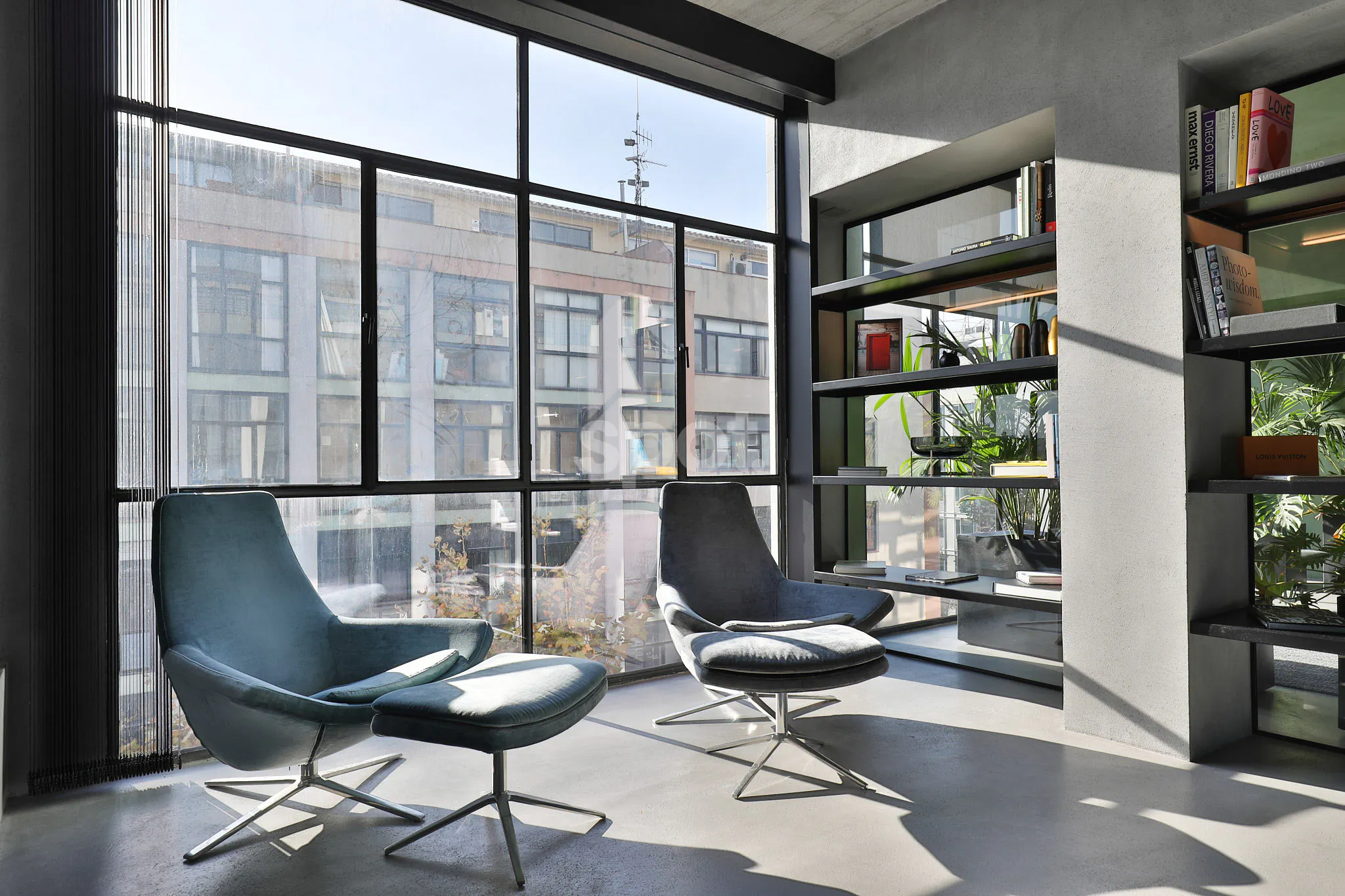 Modern grey loft filming location in Barcelona with large industrial-style windows, lounge chairs, open shelving and contemporary interior design.