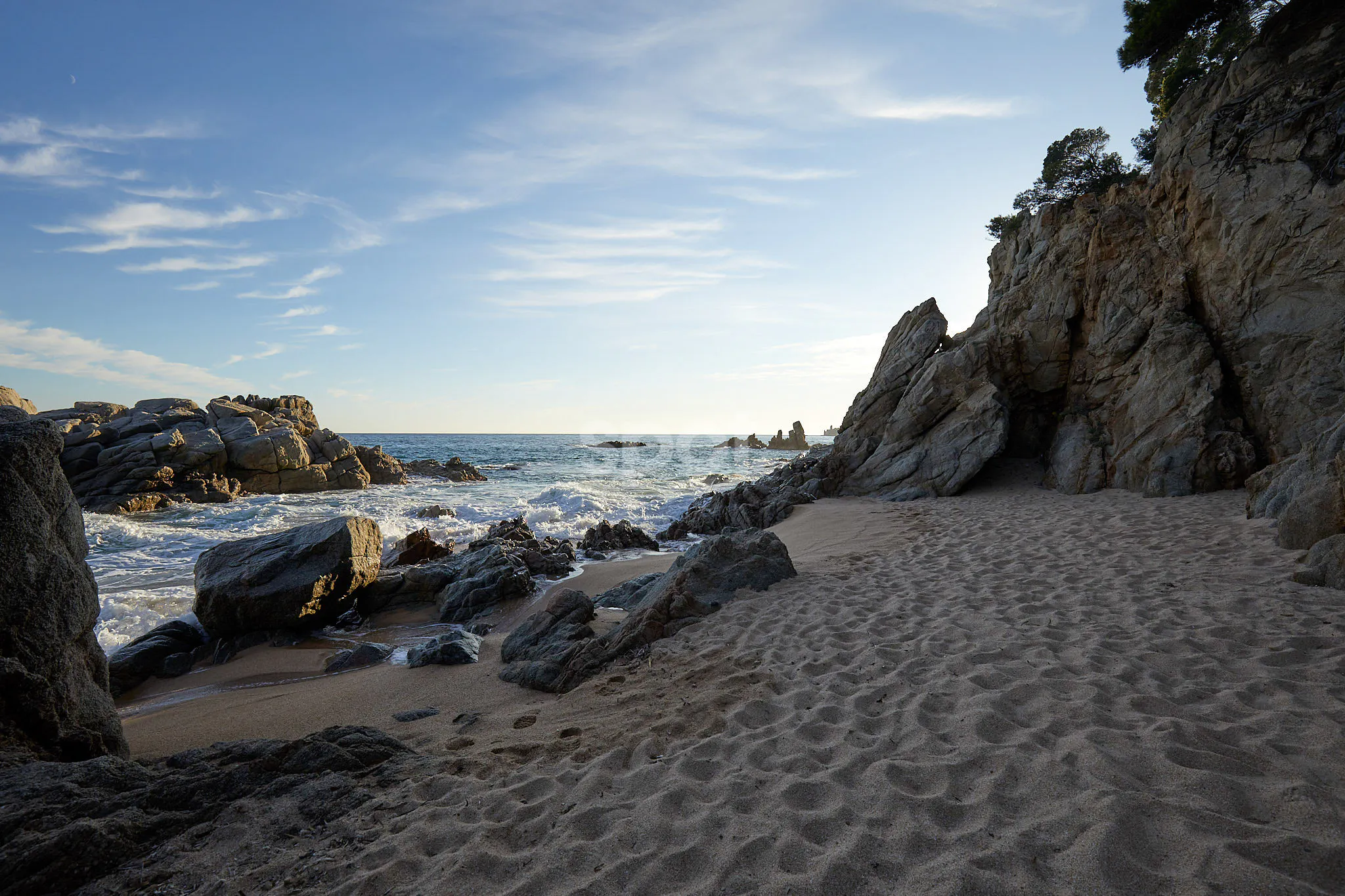 Rocky coastal cove filming location in Spain with sandy beach, sea waves, dramatic cliffs, blue sky and natural sunlight.