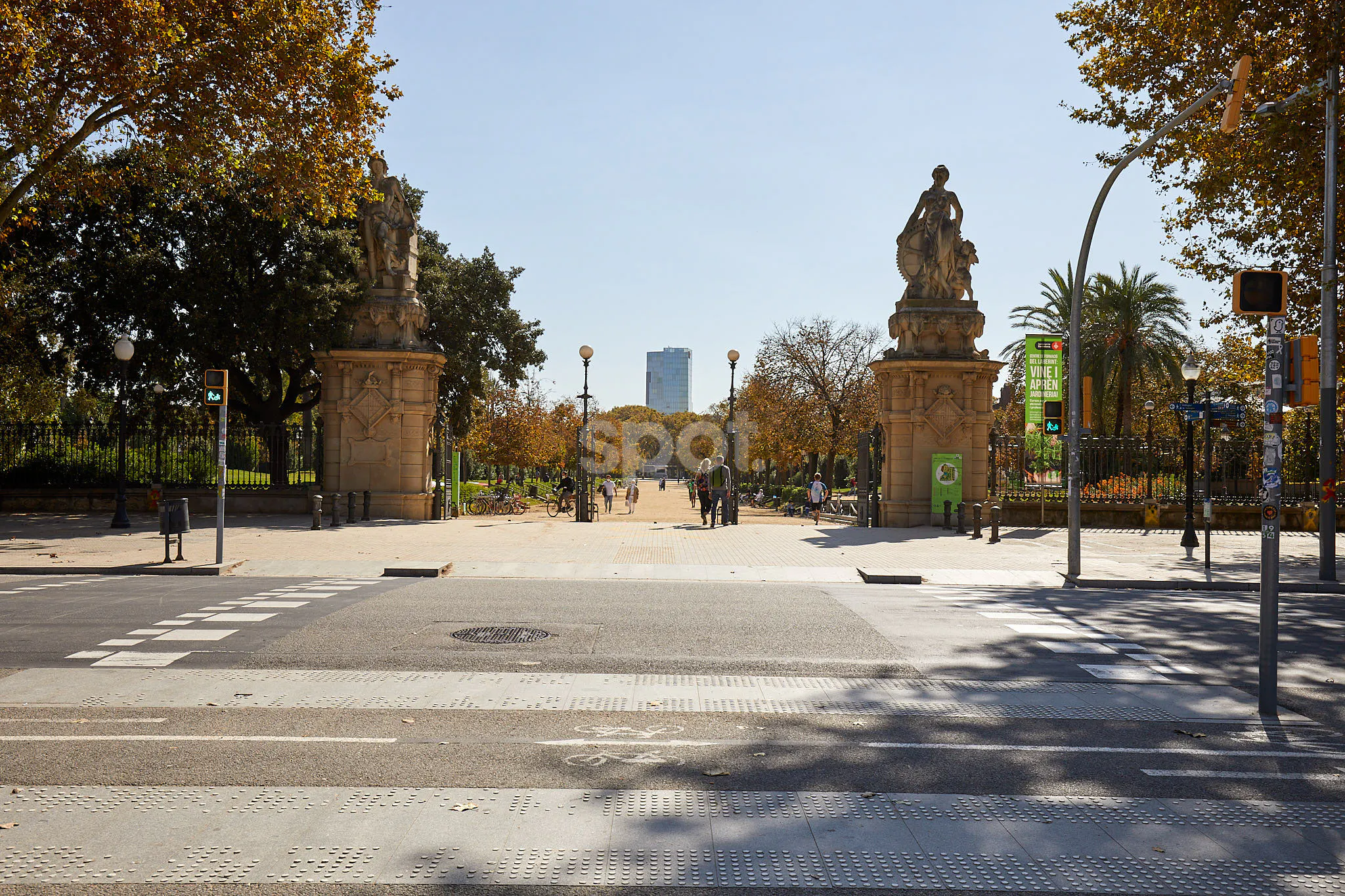 Localización de rodaje en la entrada de un parque de Barcelona con puertas históricas de piedra, esculturas, espacio público arbolado, paso de peatones y acceso urbano.