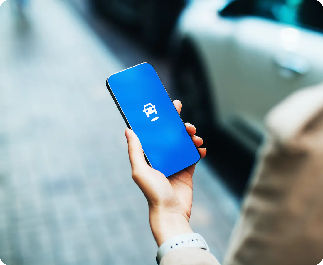A hand is holding a cell phone. On the cell phone screen is an illustration of a white car hovering above a white circle.