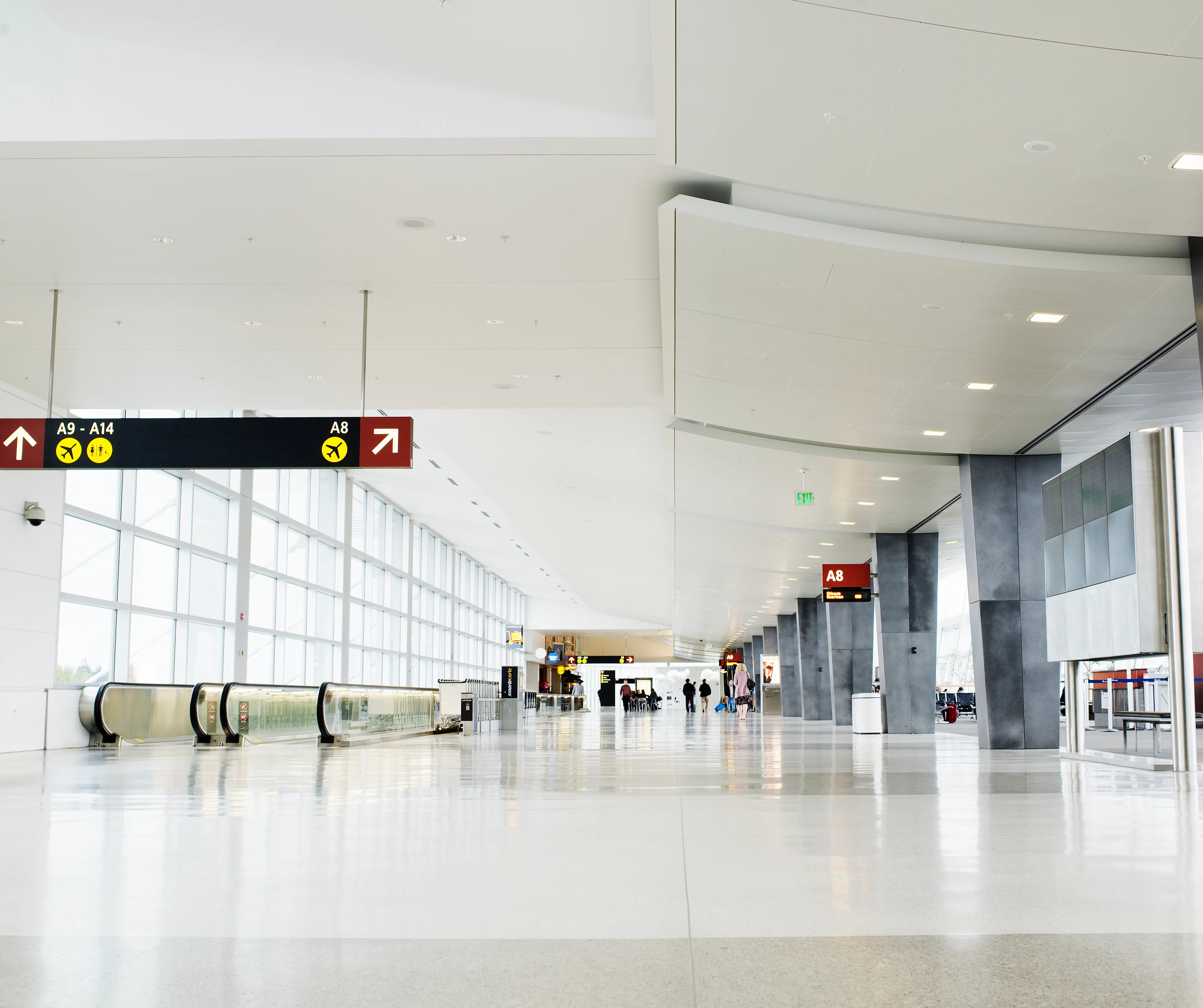a low angle shot of an empty airport concourse. there are bright, large, sunlit windows to the left with wayfaring information and escalators leading people down