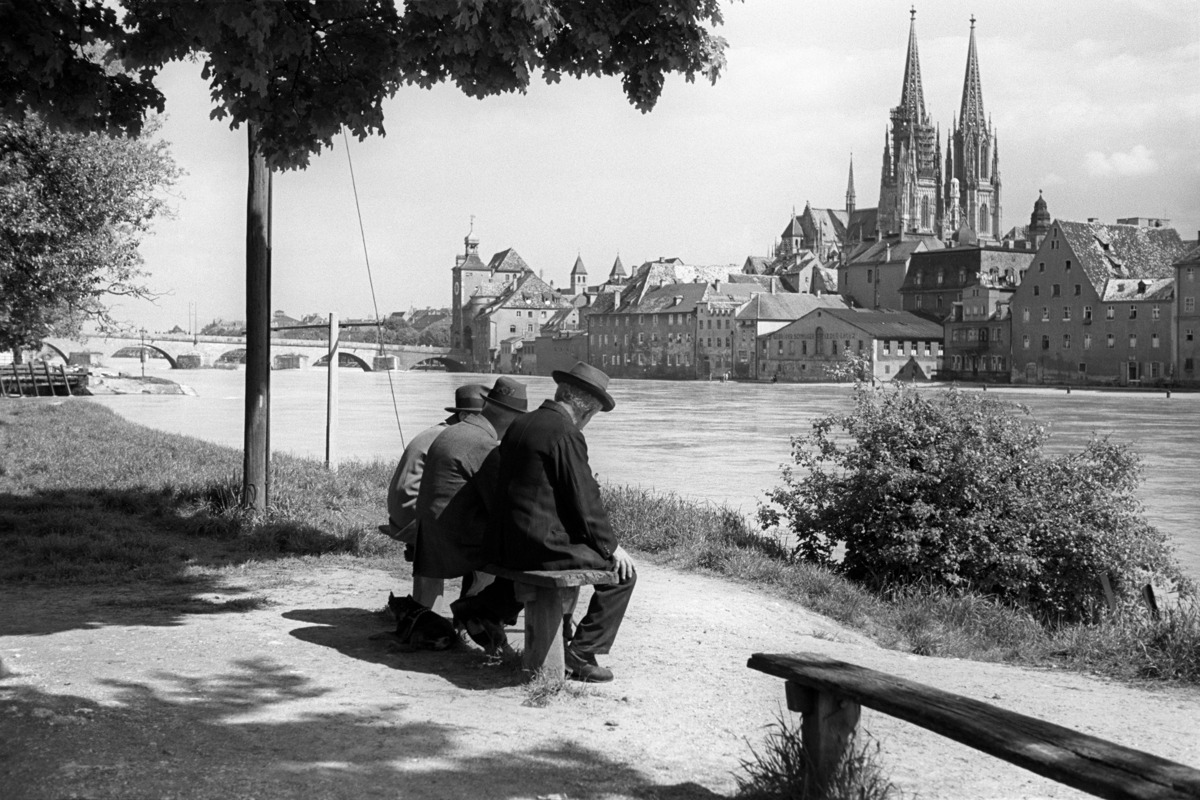 Schwarz-weiß Bild von drei Männern im Vordergrund, die auf einer Bank an der Donau sitzen. Im Hintergrund die Skyline von Regensburg.