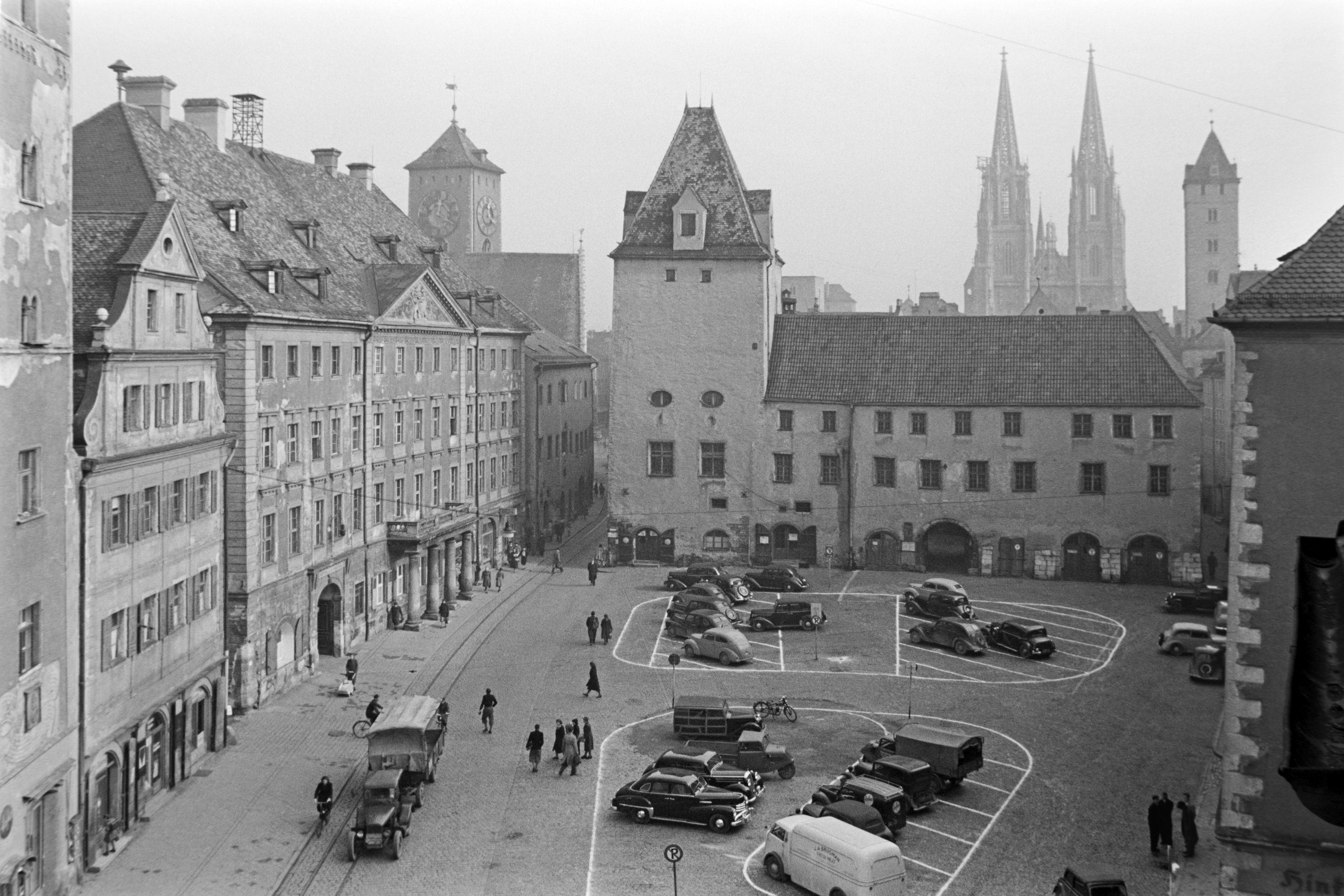 Schwarz-weiß Bild vom Haidplatz aus vergangener Zeit. Wo heute ein Brunnen und viele Freisitze von Restaurants zu finden sind, zeigt dieses Bild einen großen Autoparkplatz.