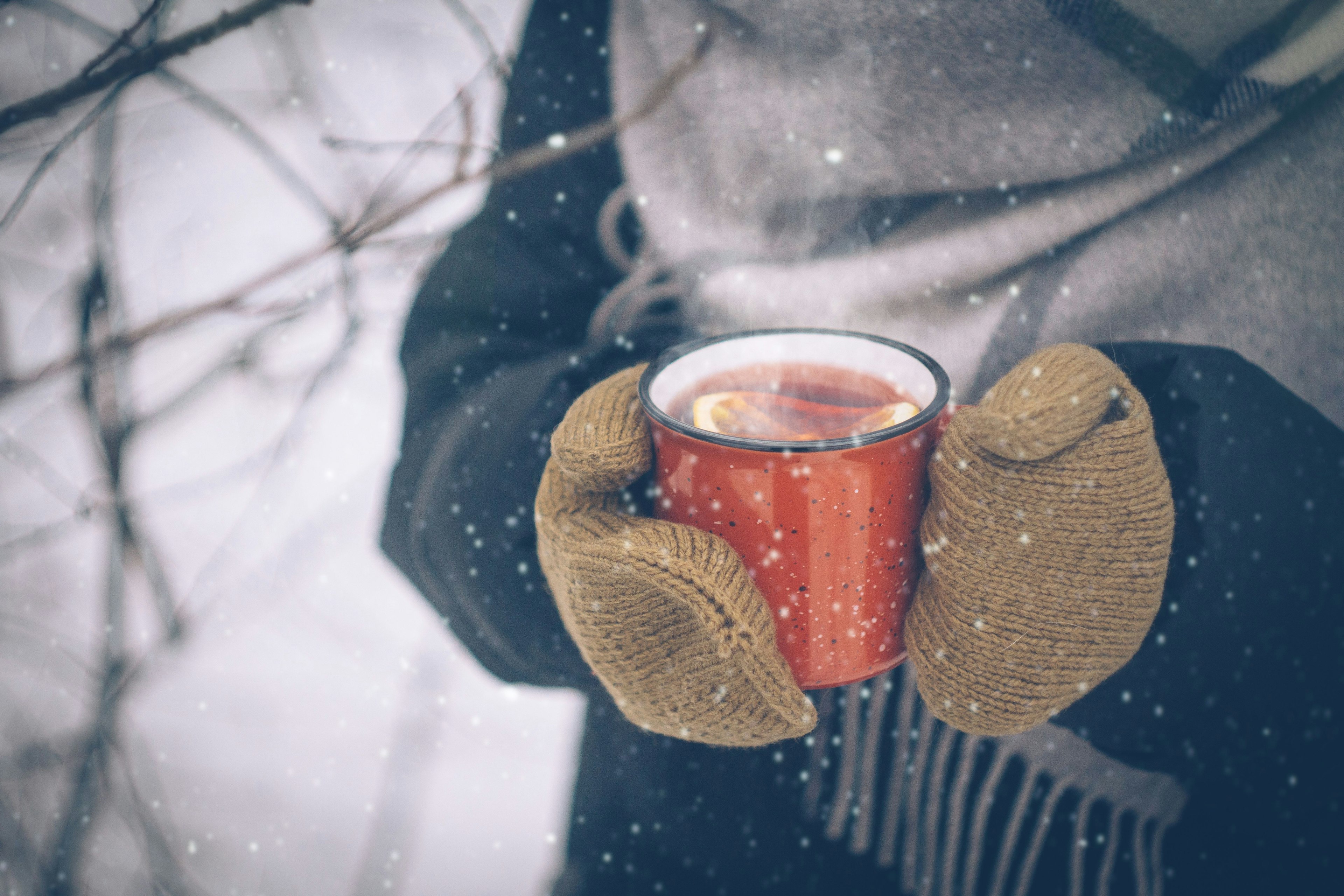 Detailaufnahme von Händen in Handschuhen, die eine Tasse mit dampfendem Inhalt in winterlicher Kulisse halten.