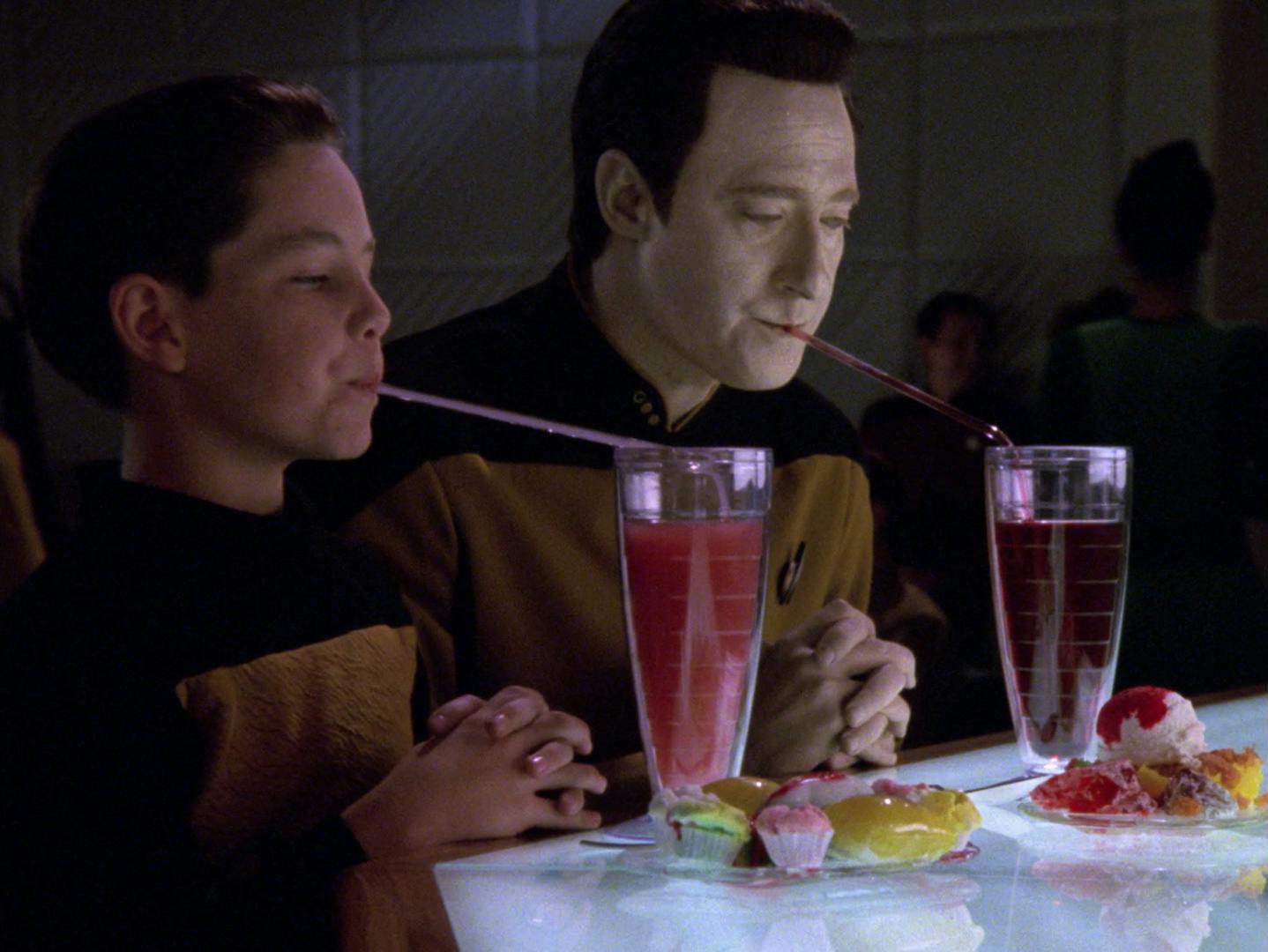 Timothy and Data sit side-by-side at 10 Forward drinking out of long straws with their hands folded and a large plate of dessert in front of them in 'Hero Worship'