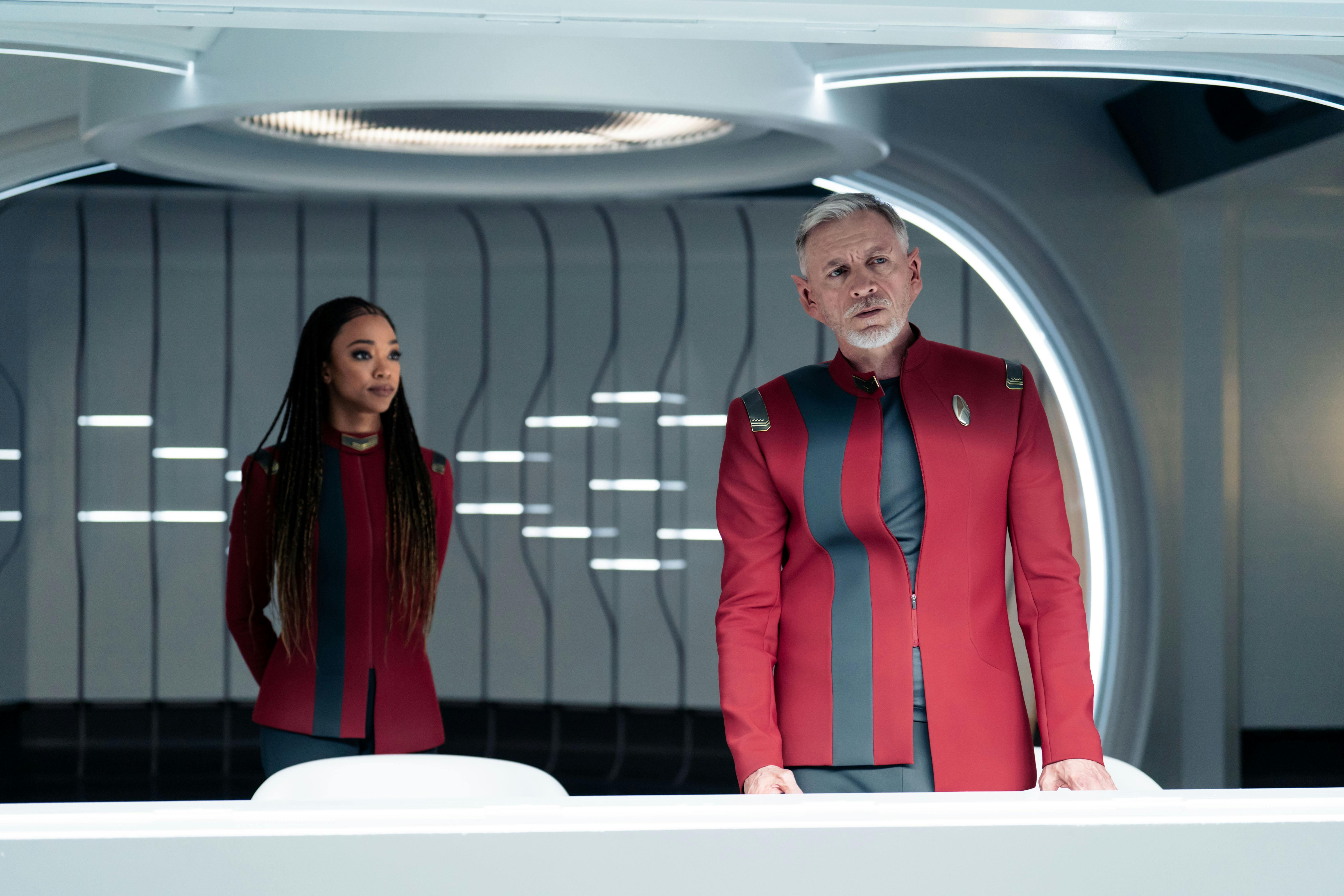 Burnham approaches Rayner, who has loosened his Starfleet uniform, as he looks out the window towards U.S.S. Discovery in 'Under the Twin Moons'