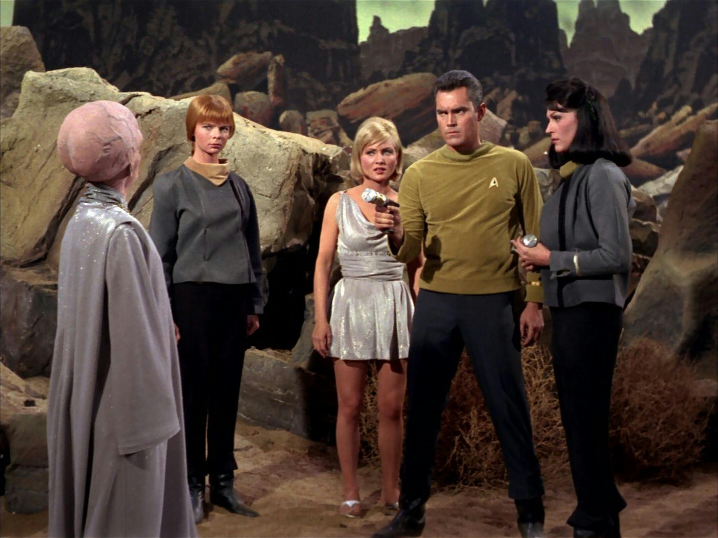 Pike points his phaser towards at the Talosian magistrate while yeoman J.M. Colt, Vina, and Number One stand by his side on Talos IV's surface in 'The Cage'