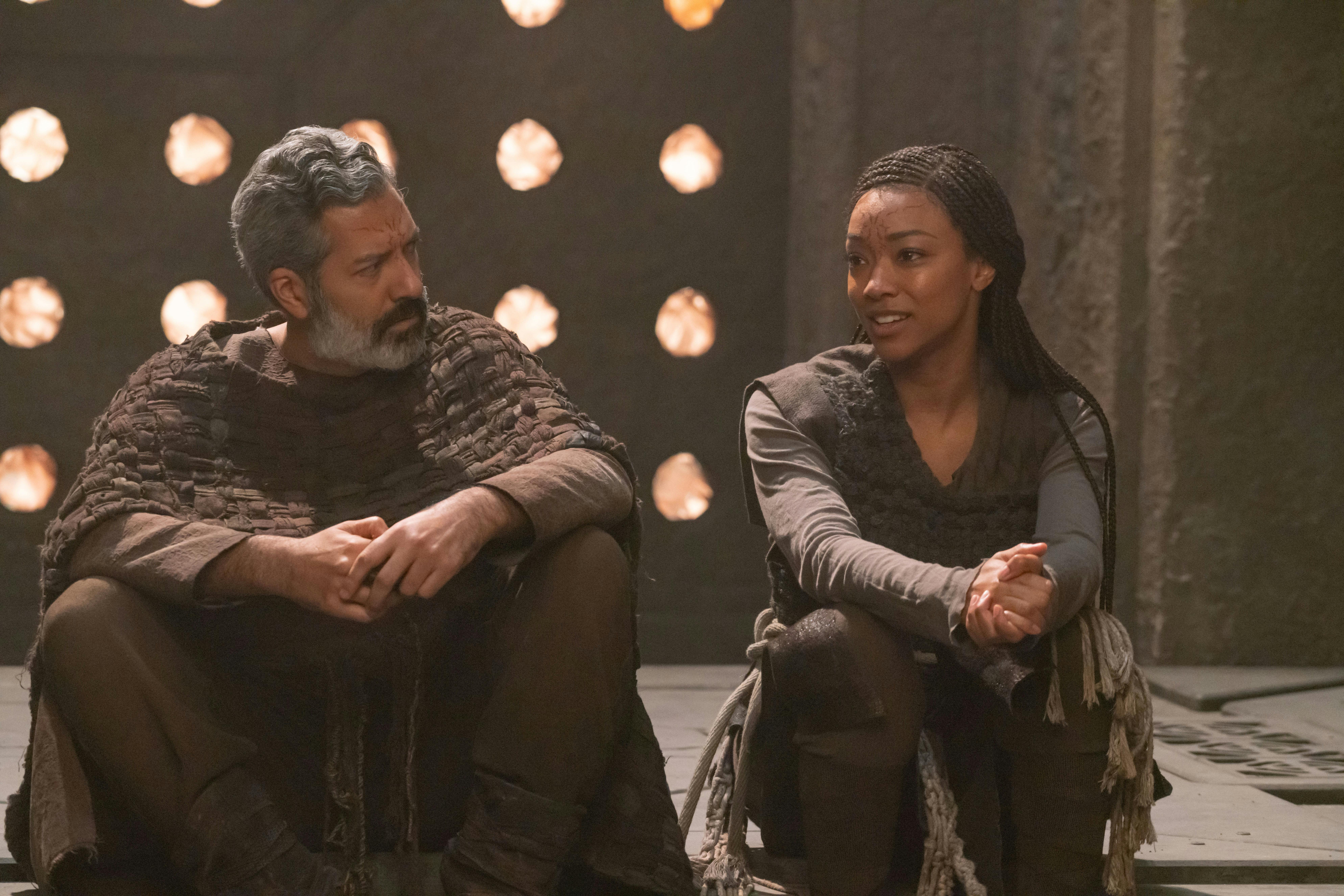 In a temple on Halem'no, Ohvahz sits next to a Michael Burnham lost in thought with her hands clasped in 'Whistlespeak'