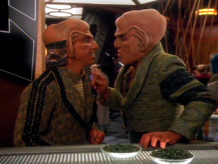 Next to the bar counter, Quark looks down while raising his finger at Rom in 'Rules of Acquisition'