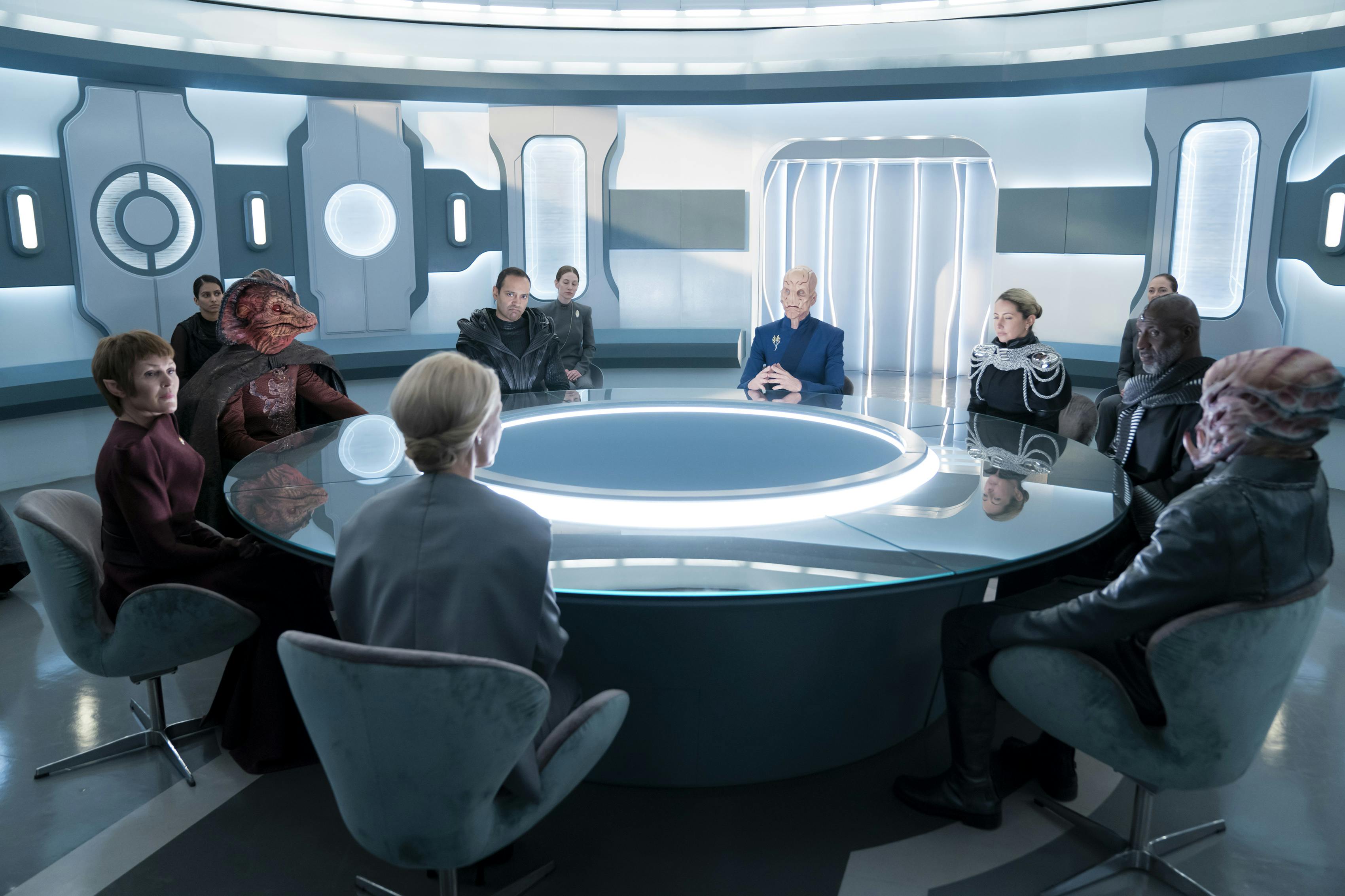 At Federation Headquarters, President T'Rina sits with Saru, a moderator, and a handful of Federation delegates around a conference table while their aides sit in the background in 'Jinaal'