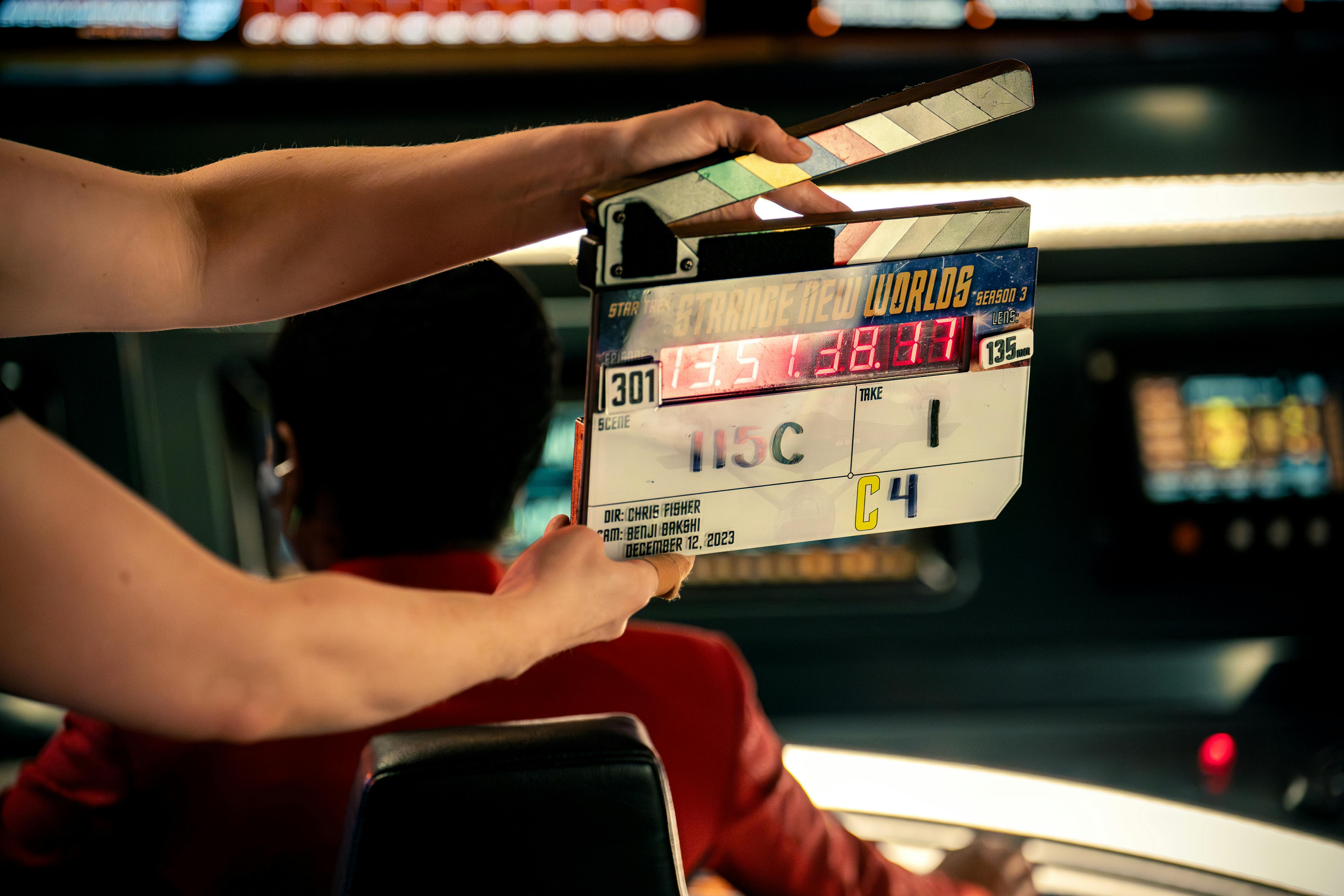 Behind-the-scenes production capture of a clapboard for Season 3, Episode 1 of Star Trek: Strange New Worlds with Celia Rose Gooding sitting at Uhura's station on the Enterprise bridge