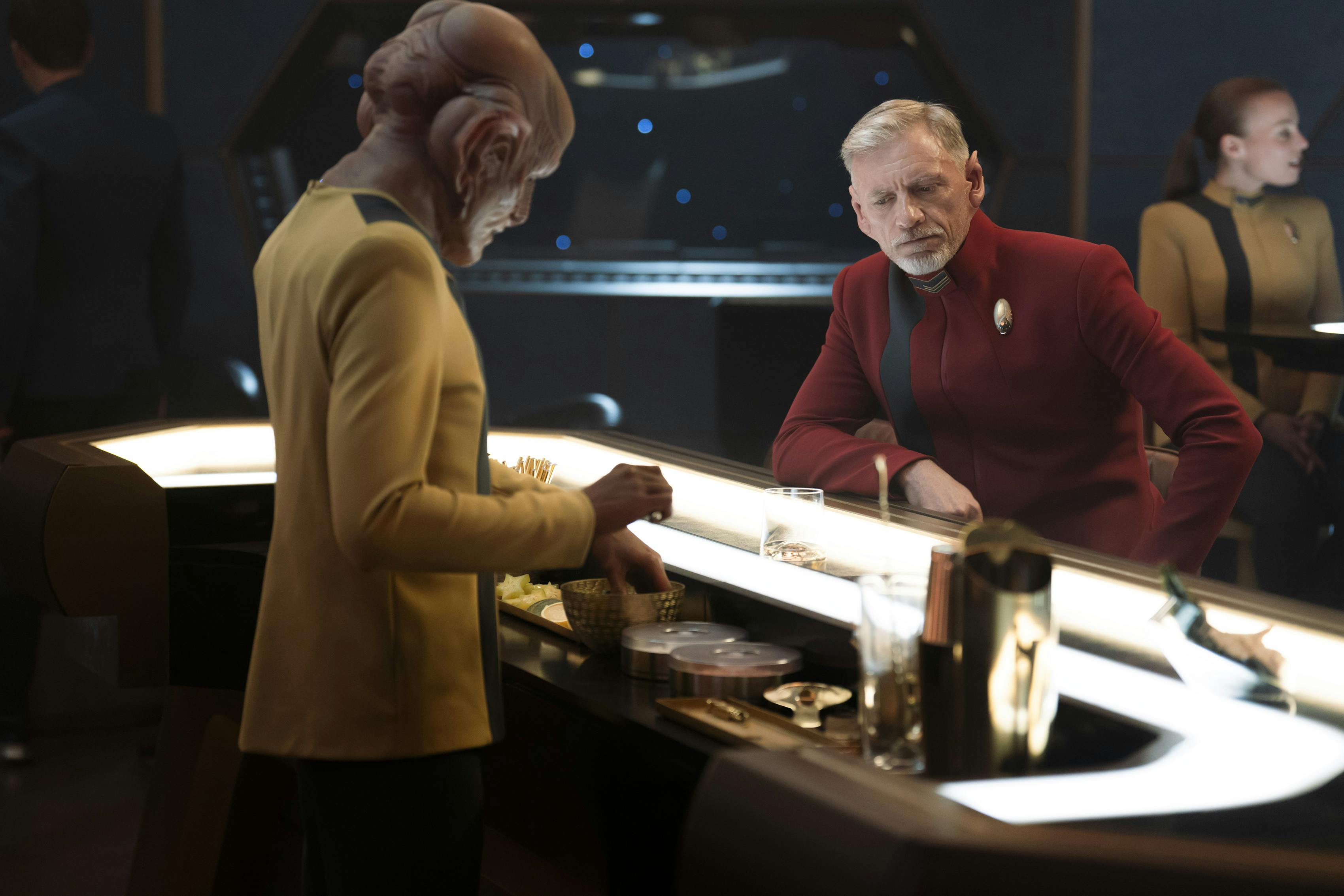 A Ferengi bartender prepares a drink for a pensive Rayner who sits at the bar in the Discovery lounge in 'Jinaal'