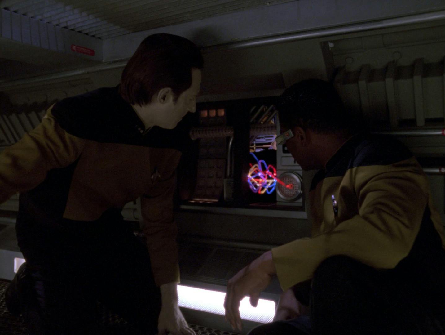 In a Jeffries tube, Data and Geordi La Forge huddle near an open panel that shows a malfunction in the ship's system in 'Emergence'