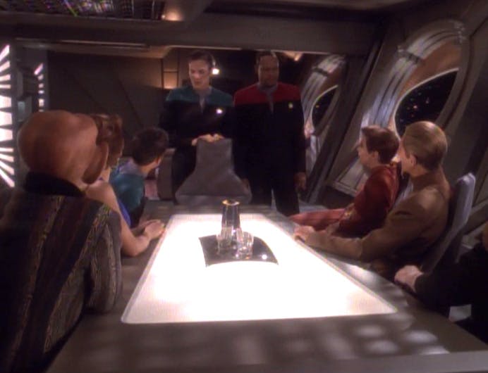 Jadzia Dax and Ben Sisko stand at the head of a conference room table surrounded by Bashir, Leeta, Quark, Kira, Odo, and O'Brien as she asks them to participate in a Trill zhian'tara ritual in 'Facets'