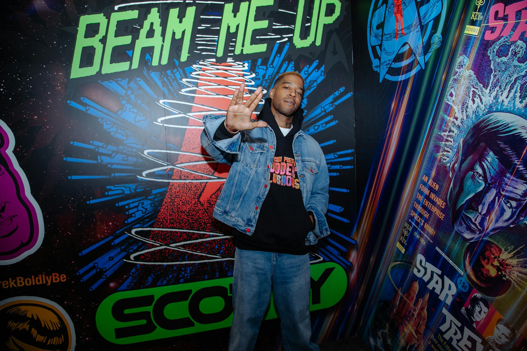 Kid Cudi standing in front of a 'Beam Me Up Scotty' mural holding up the Vulcan salute. 