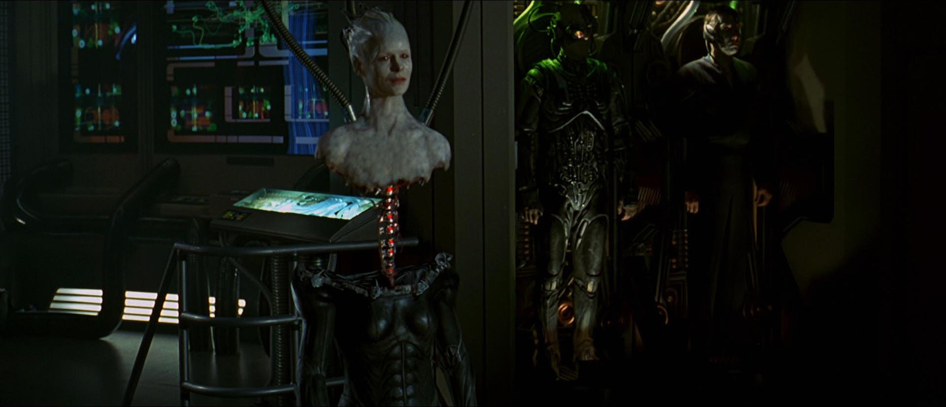 Hanging from tubing, the Borg Queen connects her vertebrae to a body as her borg hive stands behind her in 'Star Trek: First Contact' 