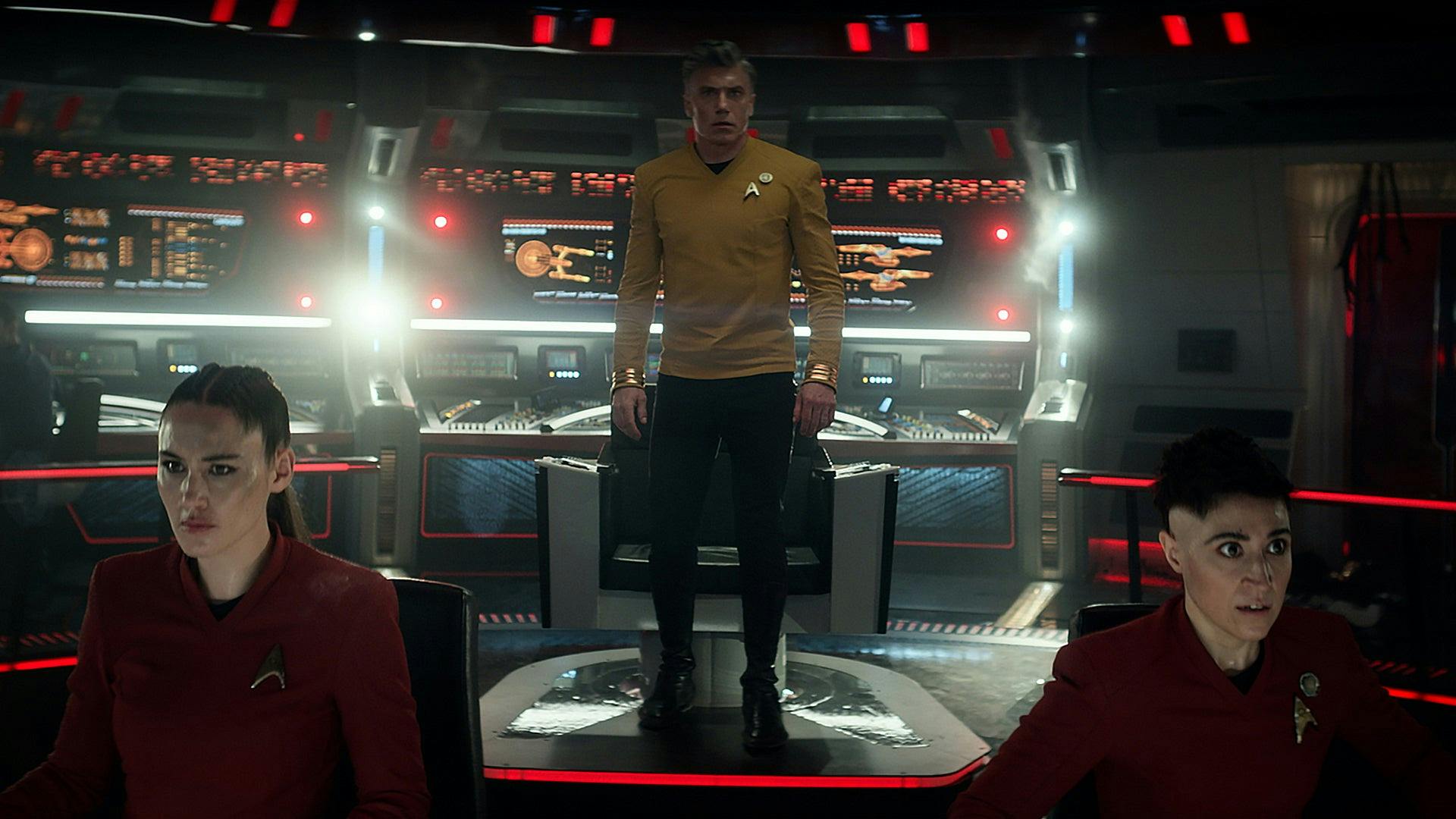 Captain Pike (Strange New Worlds) stands on the bridge in low light. At the helm, La'an Noonien Singh sits to Pike's right and Erica Ortegas sits to Pike's left.