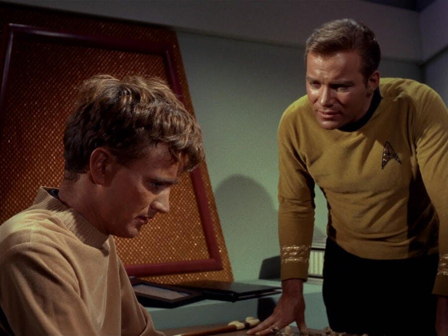 Captain Kirk (The Original Series) leans forward with his hands resting on a table as he addresses Charlie from the episode 