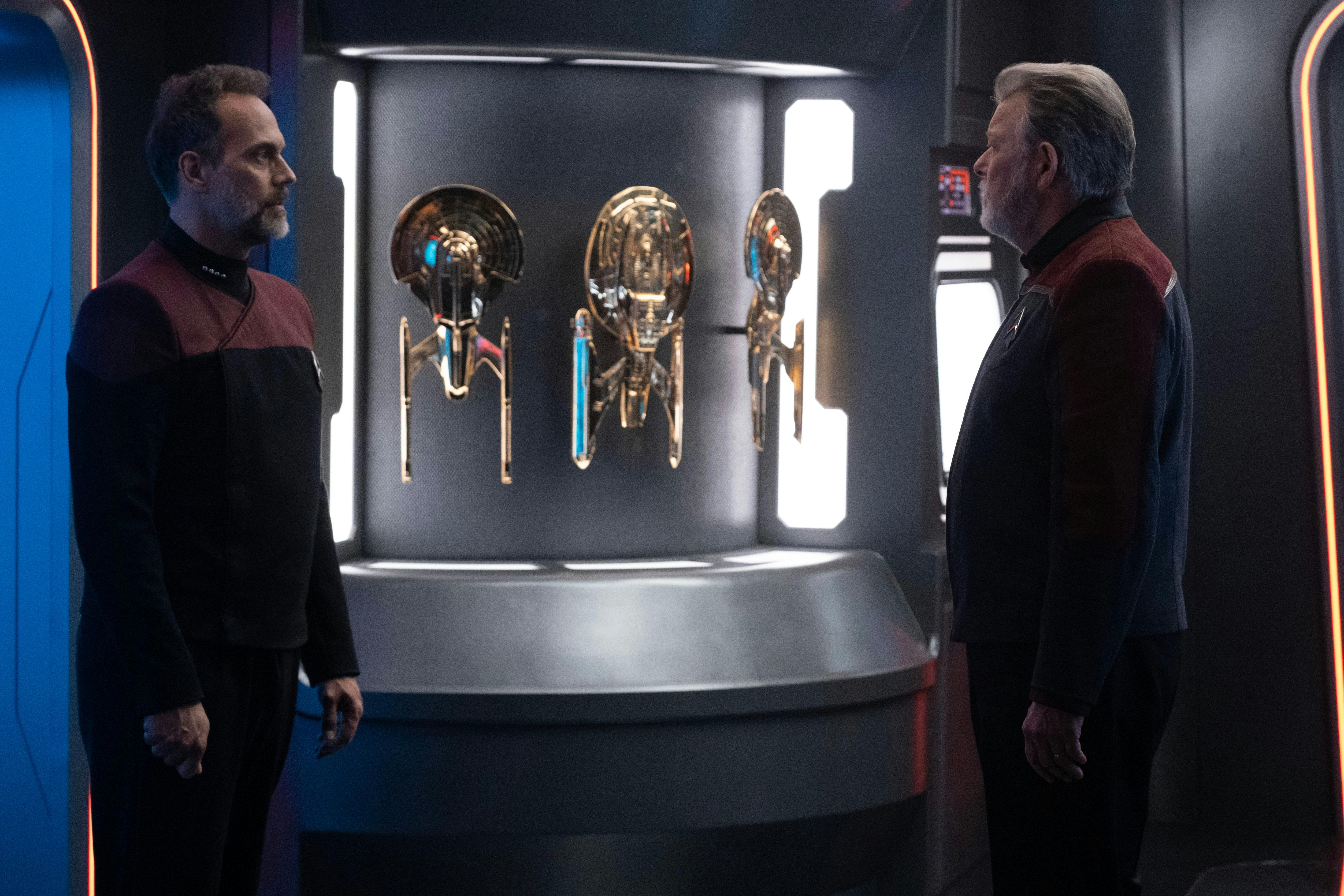 Captain Liam Shaw and Captain Will Riker stand face-to-face in front of a display of gold-plated starships on Star Trek: Picard