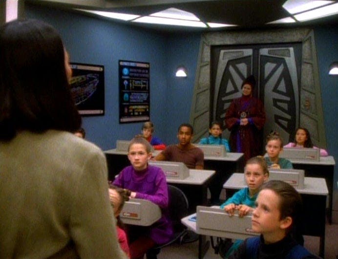 Keiko O'Brien stands before her classroom aboard Deep Space 9 as Winn Adani stands in the back observing her with her hands clasped in front of her in Star Trek: Deep Space Nine's 'In the Hands of the Prophets'
