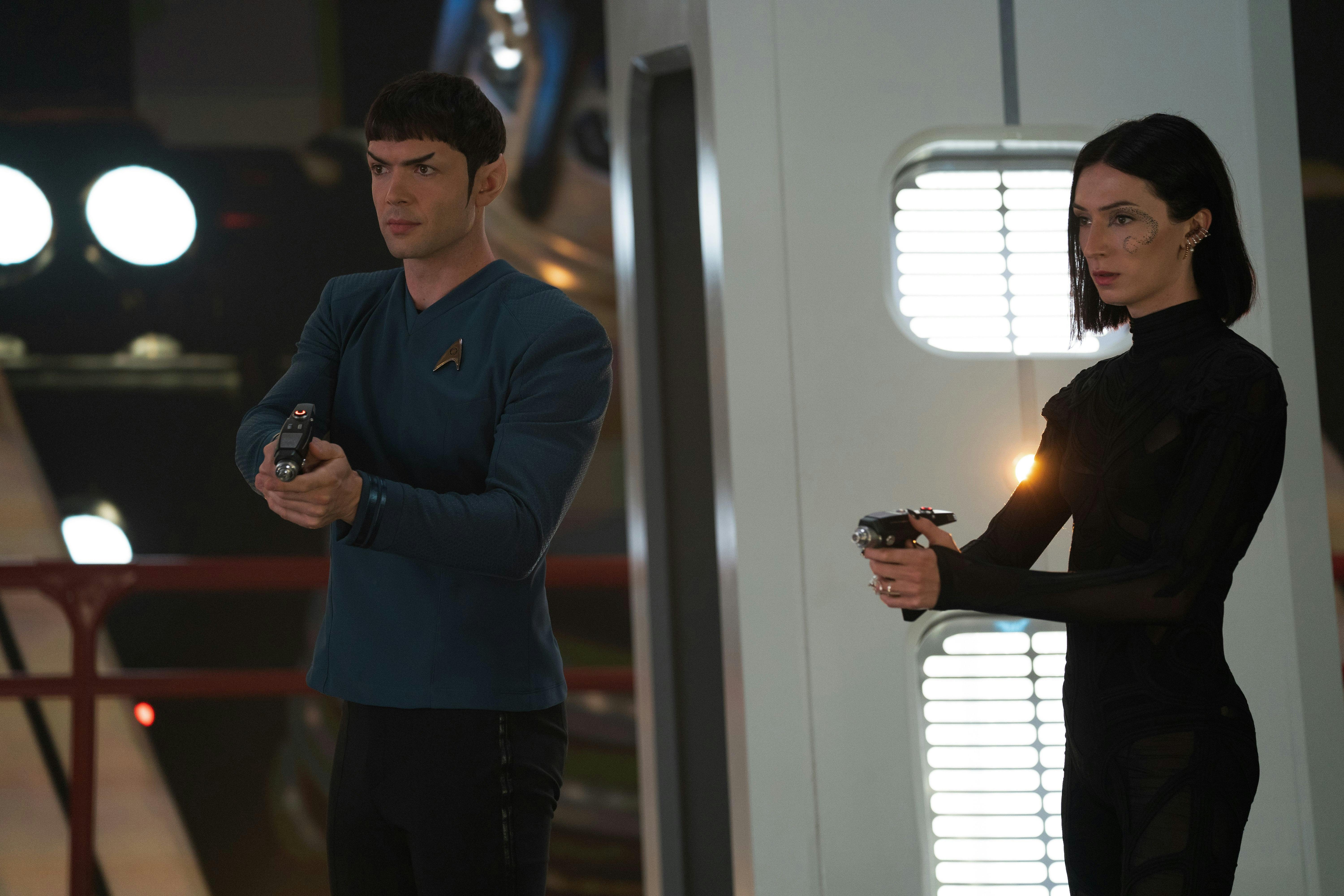 Spock (Ethan Peck) stands next to Dr. Aspen (Jesse Jame Keitel). Both characters are holding phasers.