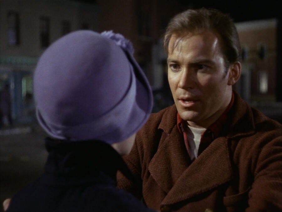 Captain Kirk (The Original Series), dressed in 20th century clothes, talks to Edith Keeler while holding her arms.