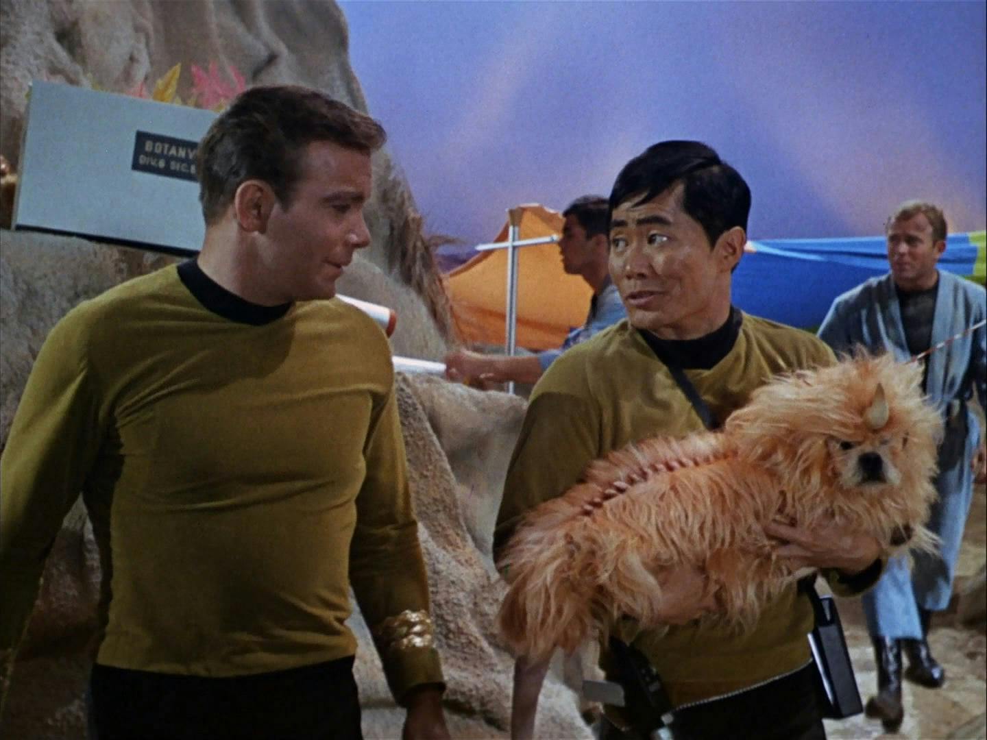 Captain Kirk and Sulu (The Original Series) stand on a planet. Sulu is holding an alien dog in his arms. Both men are smiling.