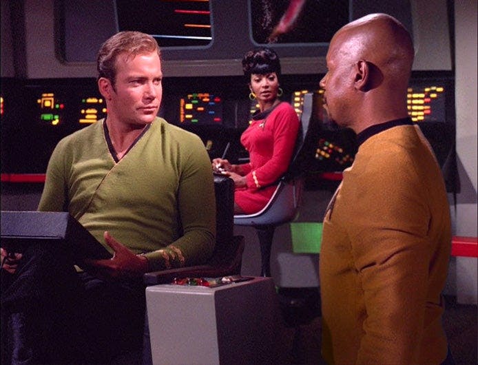 Captain Sisko talks to Captain Kirk on the bridge of the Enterprise. Uhura looks on in the background.