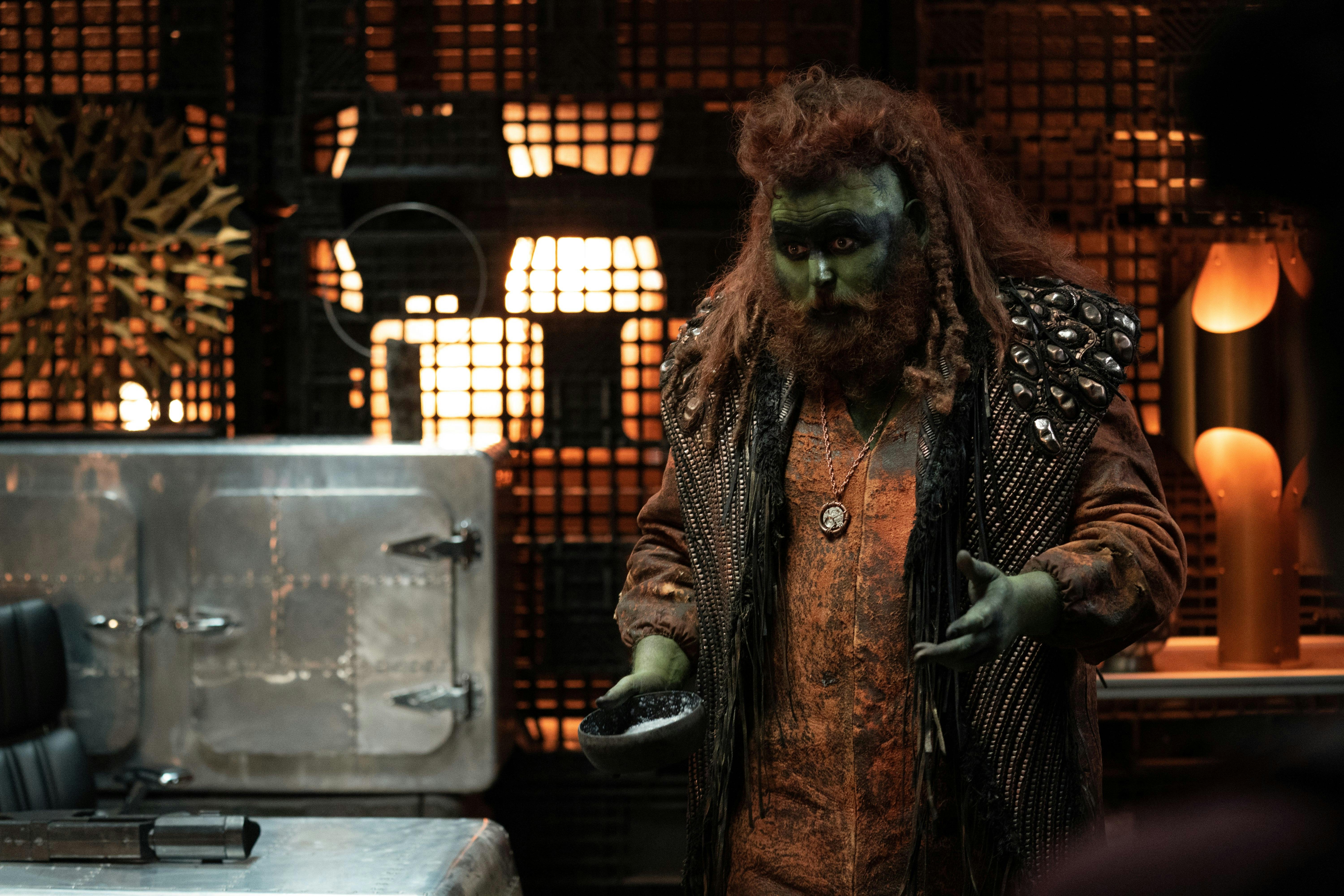 A green alien with red hair and a red beard stands talking to someone off camera. 