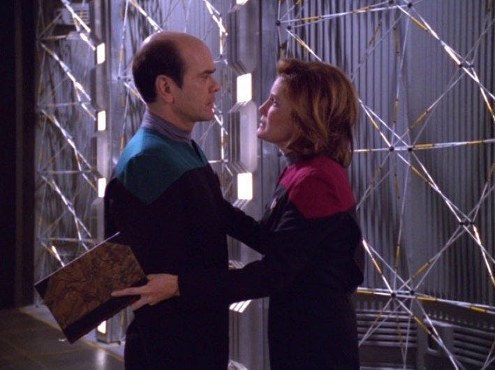 While still clasping an open book, Kathryn Janeway places her hands on both of The Doctor's arms in 'Latent Image'