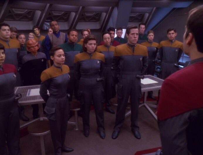 Crew members of the U.S.S. Valiant, including Nog and Jake Sisko, listening to Acting Captain Tim Watters in the mess hall in 'Valiant'