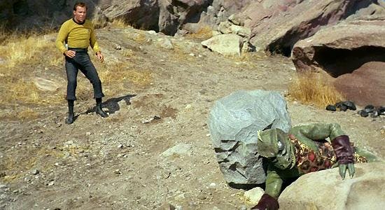 Kirk stands in the background as the Gorn is trapped between two boulders in the foreground of Star Trek: The Original Series' Arena