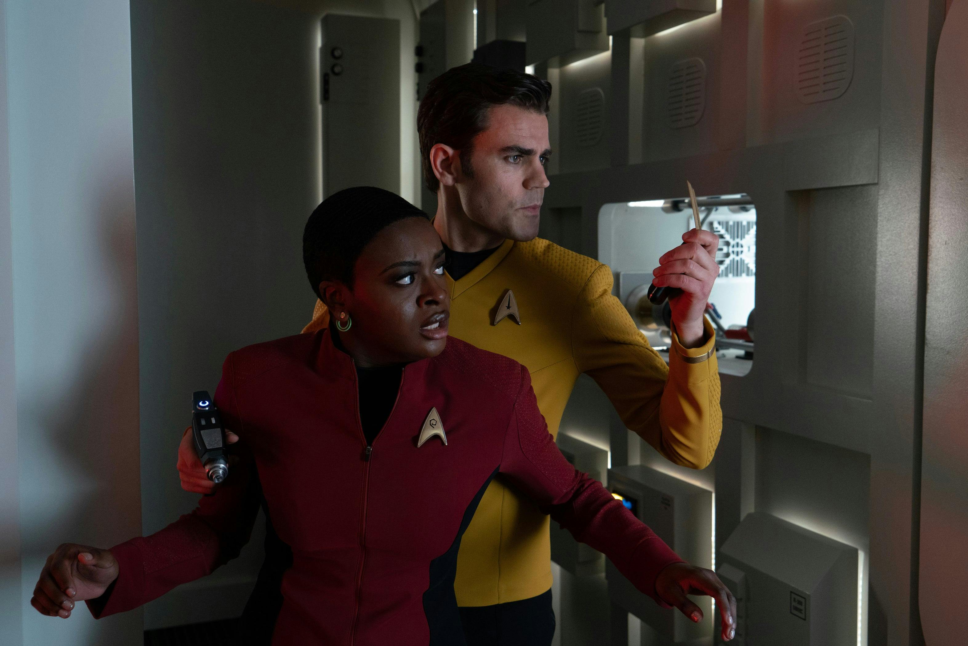 James Kirk stands looking at the tricorder in his left hand while holding his phaser in his right hand leaning against Uhura's right arm as she stands in front of him with her back to him in 'Lost In Translation'