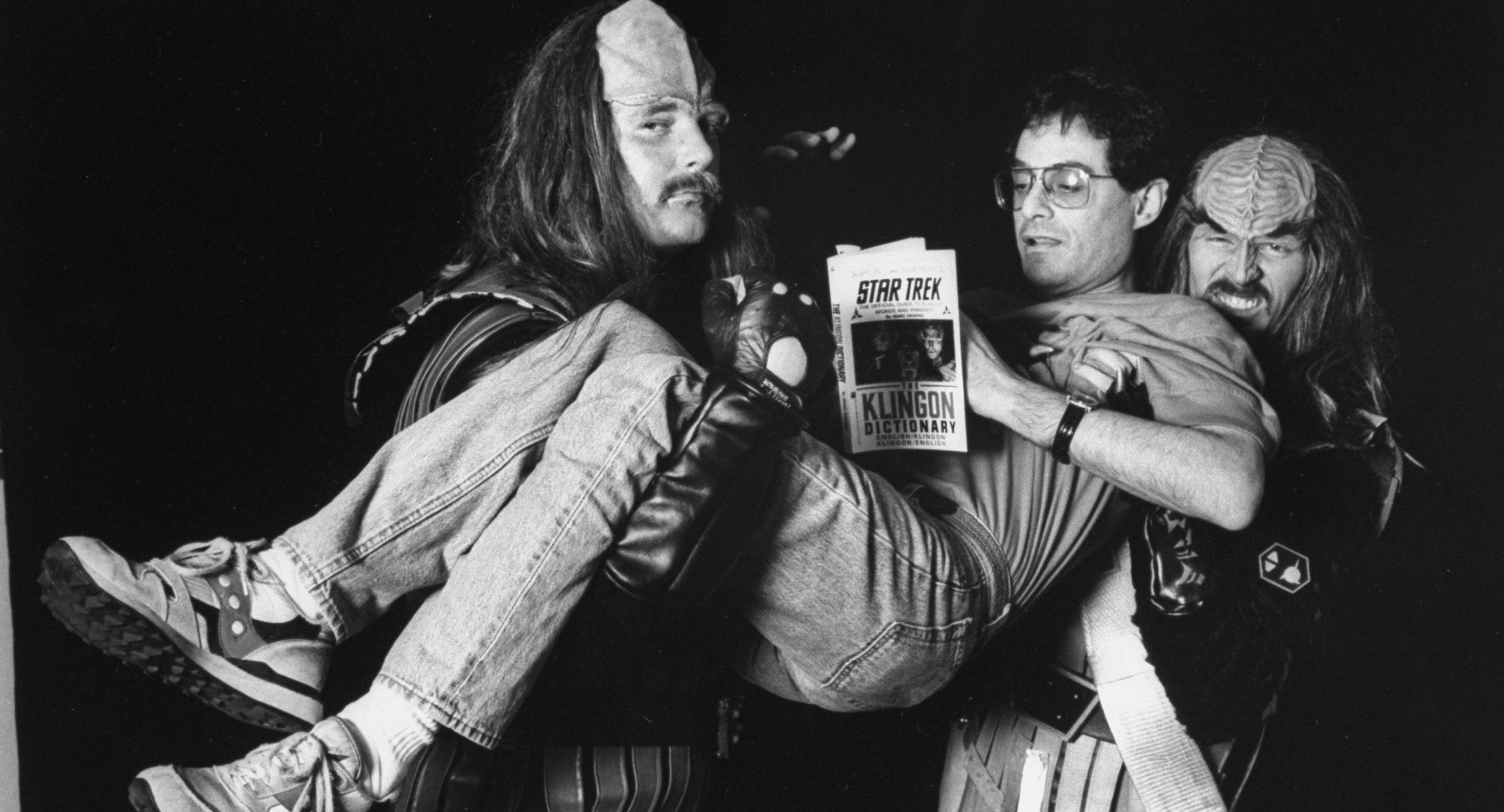 Two men in Klingon makeup and costume lift up Marc Okrand as he flips through his book 'The Klingon Dictionary'