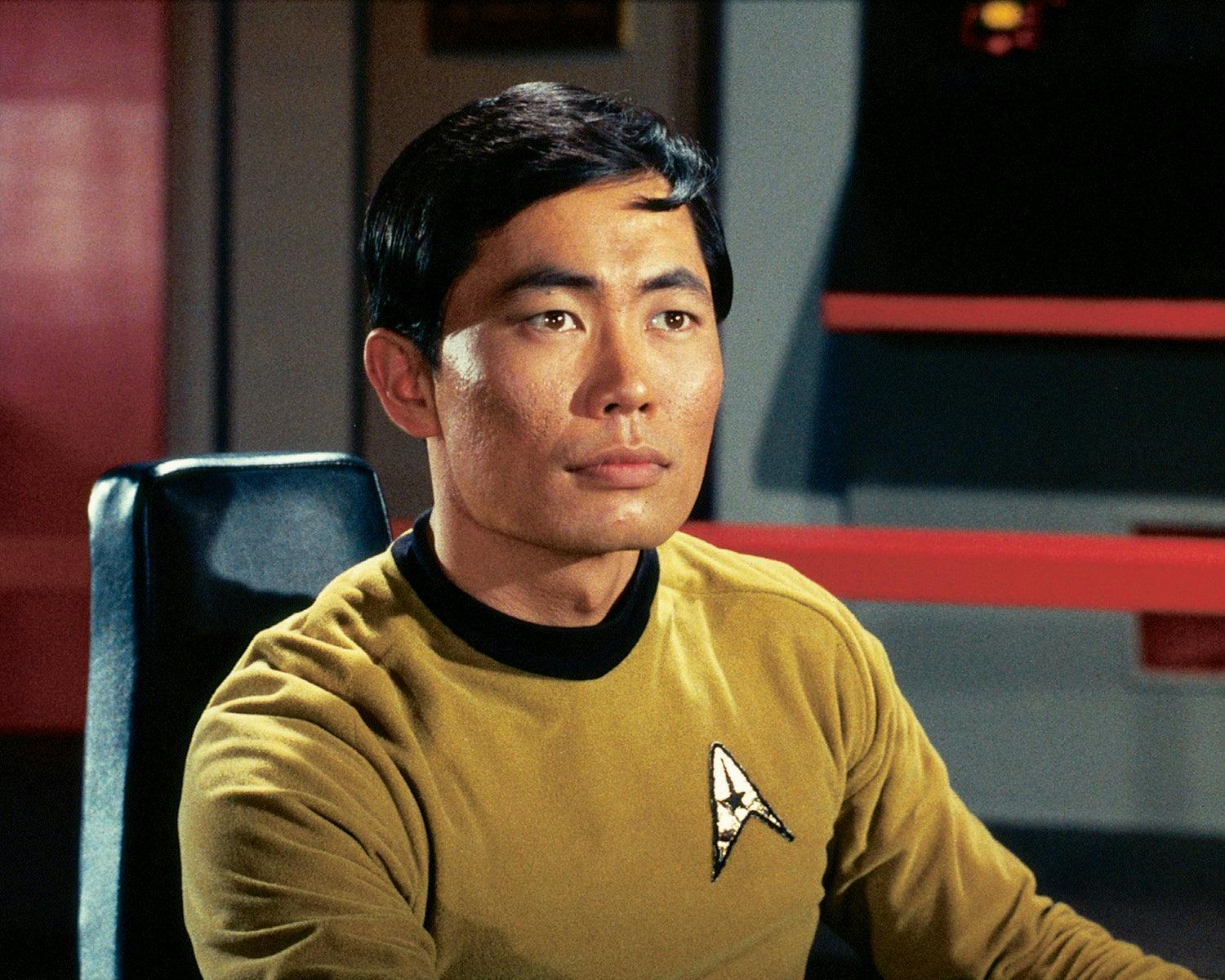 A publicity photo of George Takei as Hikaru Sulu, wearing a gold shirt and sitting at the helm of the Enterprise.