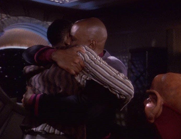 Captain Sisko embraces his son, Jake, as Nog watches.