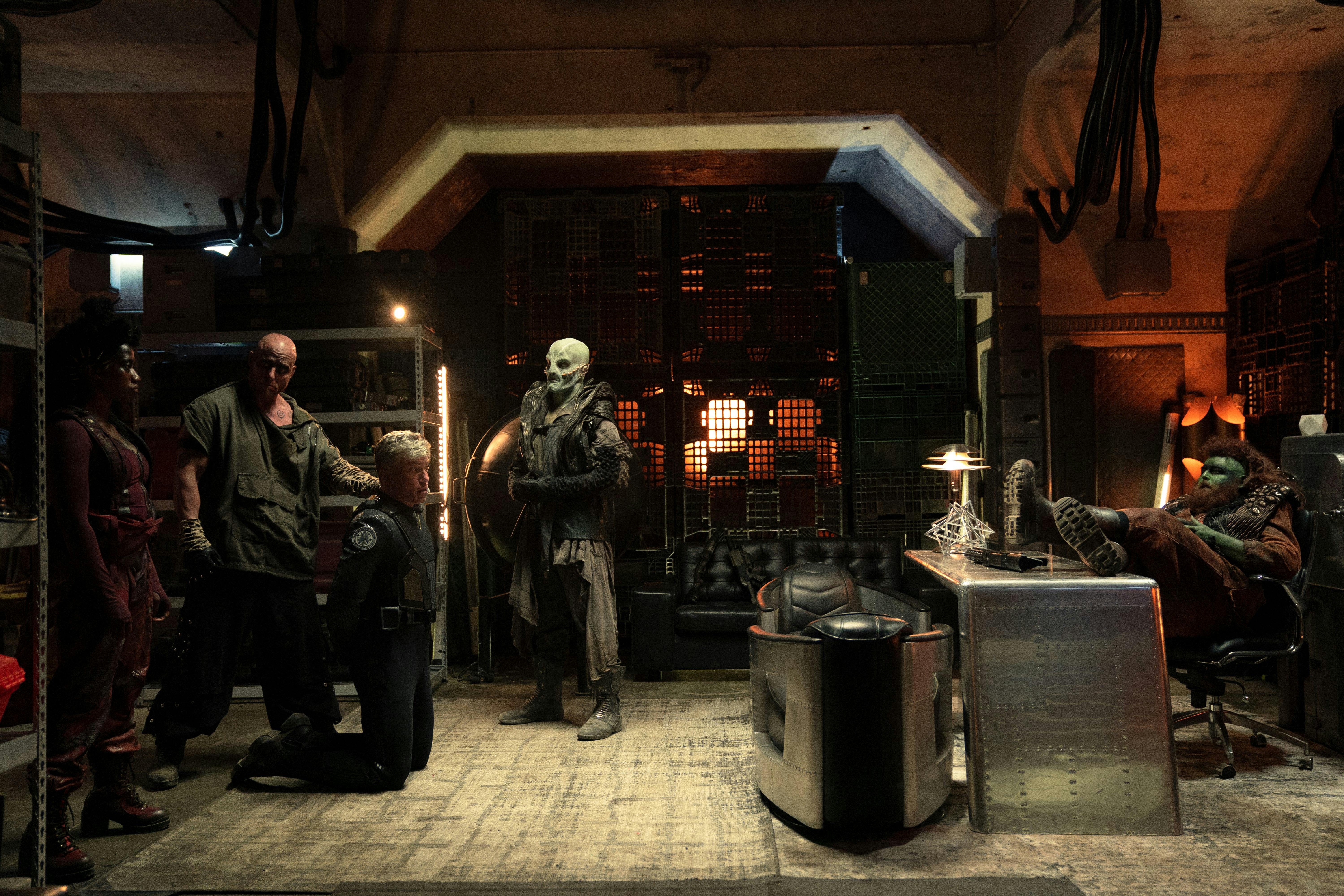 An alien with green skin and an orange beard and hair sits at a desk. Across the room, Pike (Anson Mount) is kneeling next to two alien guards.