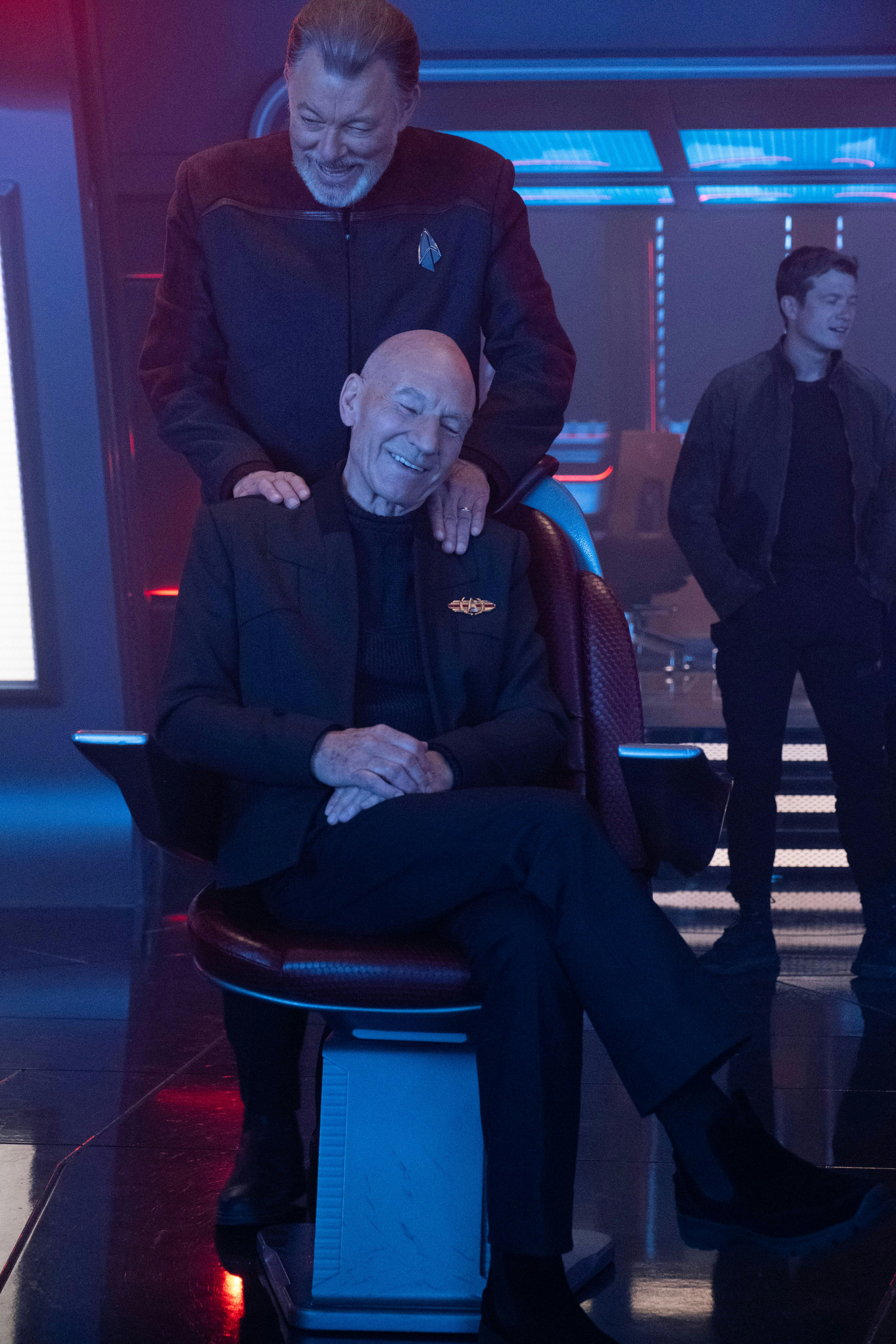 Star Trek: Picard BTS still - Patrick Stewart sits in the captain's chair smiling while Jonathan Frakes stands behind with his hands on his shoulder