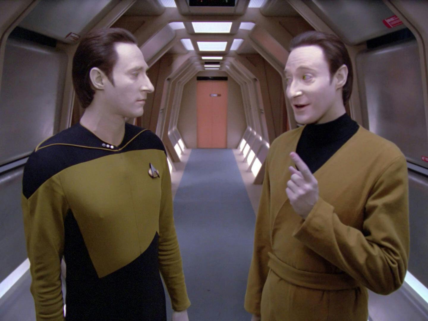 Data and Lore stand in one of the hallways on the Enterprise-D. Data stands to the left, and Lore is on the right. Lore is gesturing and smiling, while Data has a neutral expression.