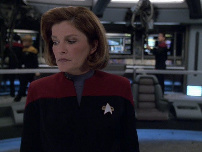 Captain Janeway stands on the bridge, looking thoughtful.