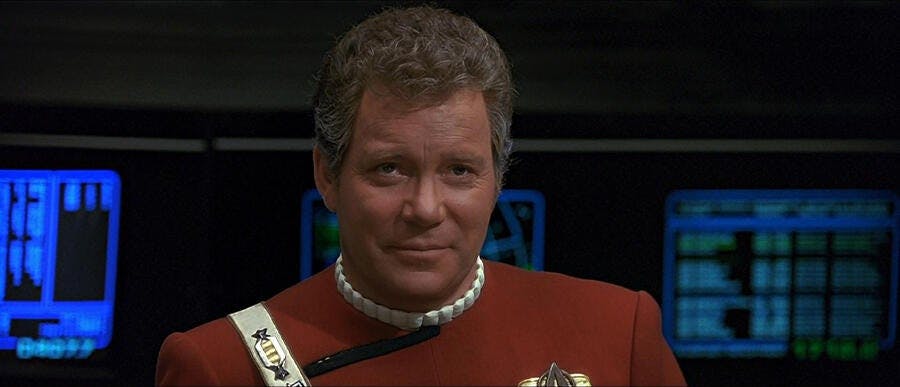 Captain Kirk (The Original Series), wearing the red uniform of the TOS movies, smiles at the viewscreen on the bridge.