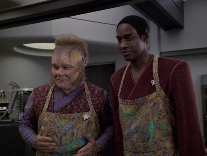 Neelix and Tuvok stand next to each other. Both are wearing aprons, and both are smiling in 'Riddles'